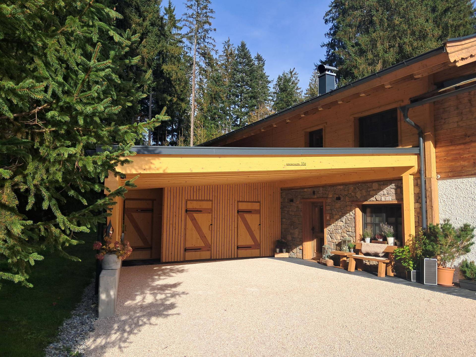 Holzhaus mit Carport und Kiesauffahrt, umgeben von Bäumen an einem sonnigen Tag.