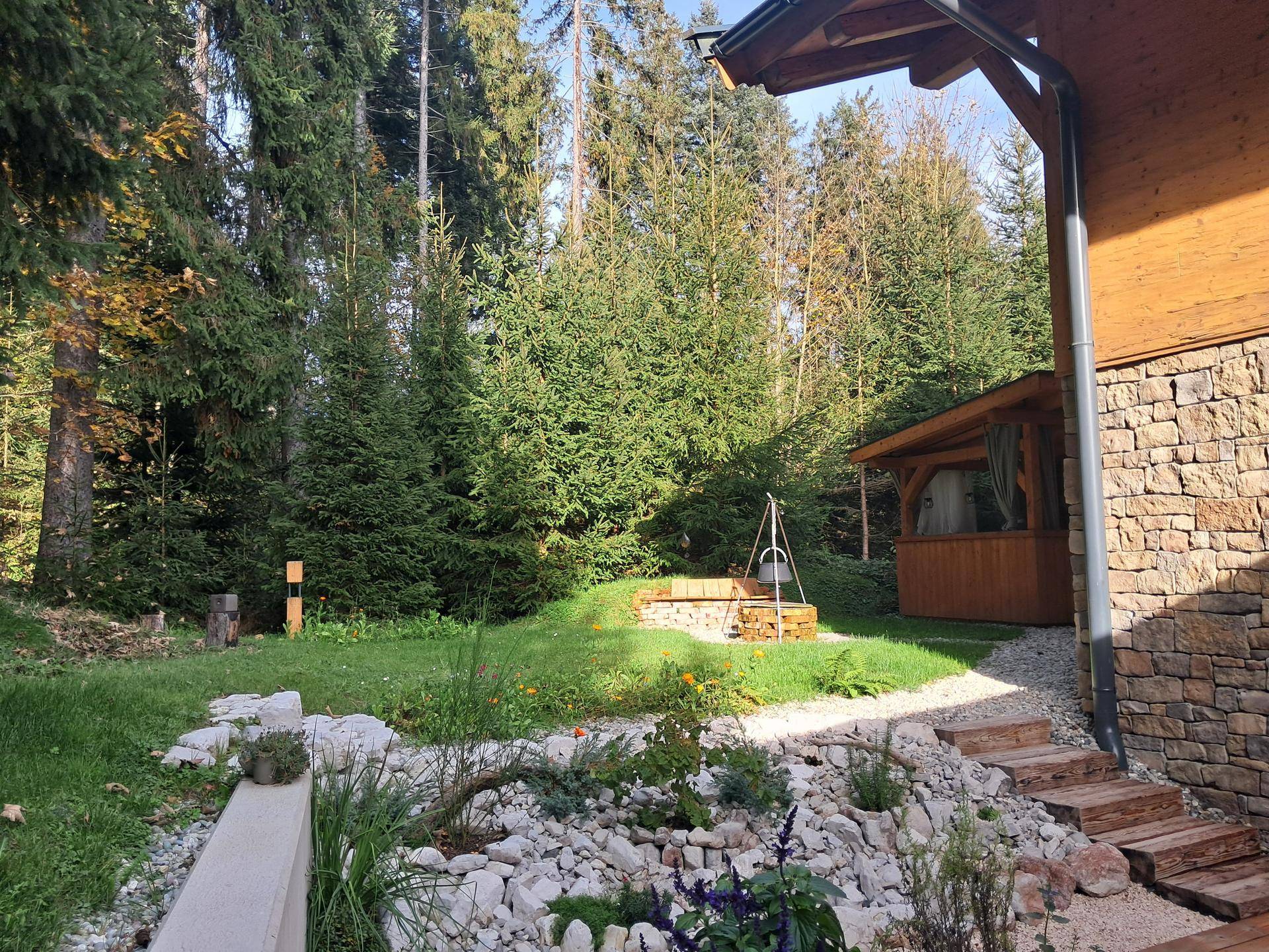 Naturnaher Gartenbereich mit Feuerstelle, Holzpavillon und umgebendem Wald.