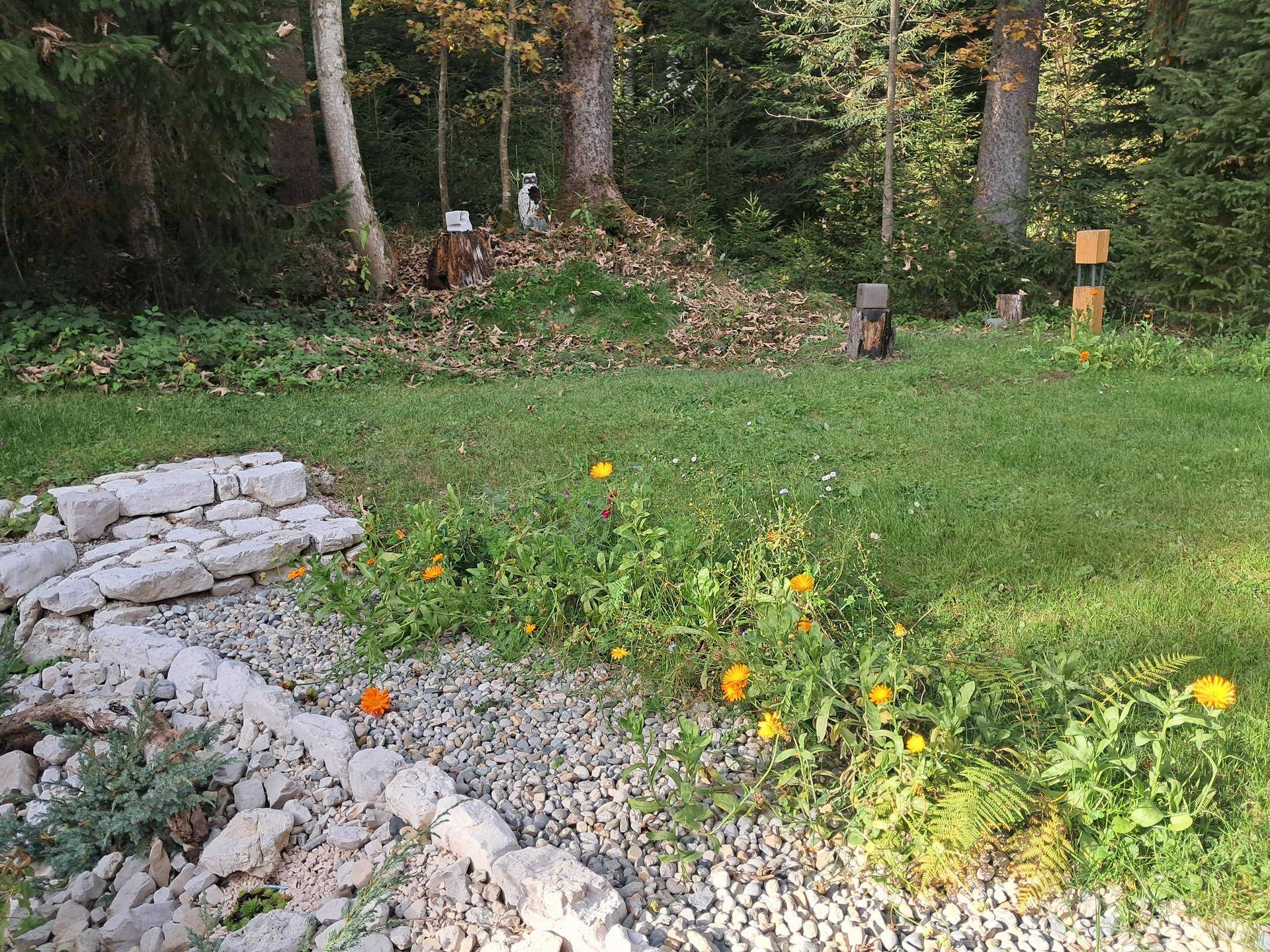 Gartenabschnitt mit Blumenbeeten, Kieswegen und einer Steinmauer am Waldrand.