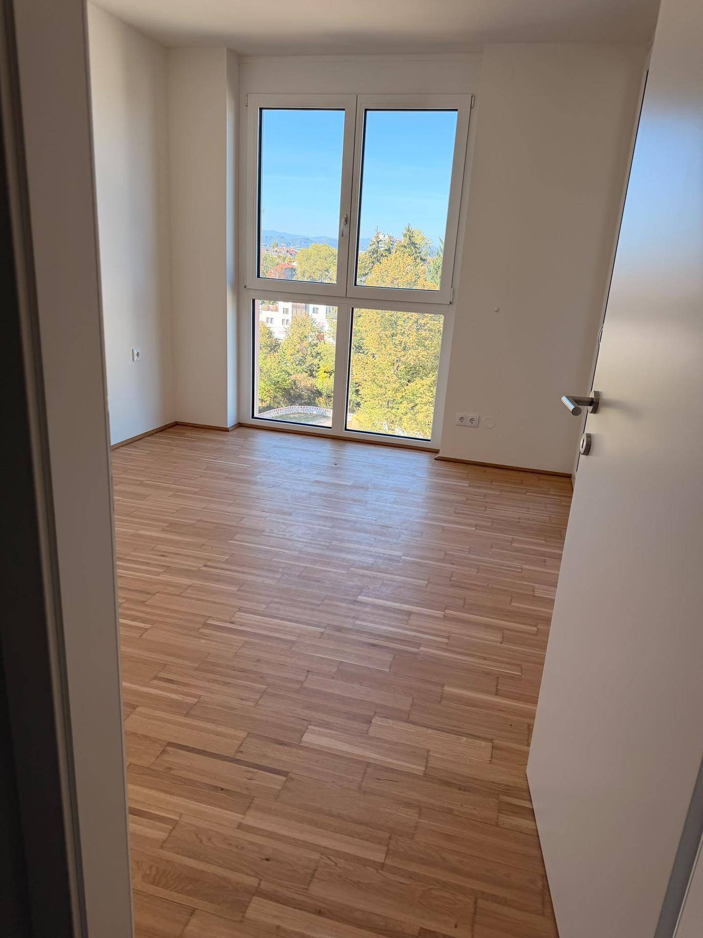 Leeres Zimmer mit hellem Holzboden und großem Fenster mit Blick ins Grüne und auf die Ferne.