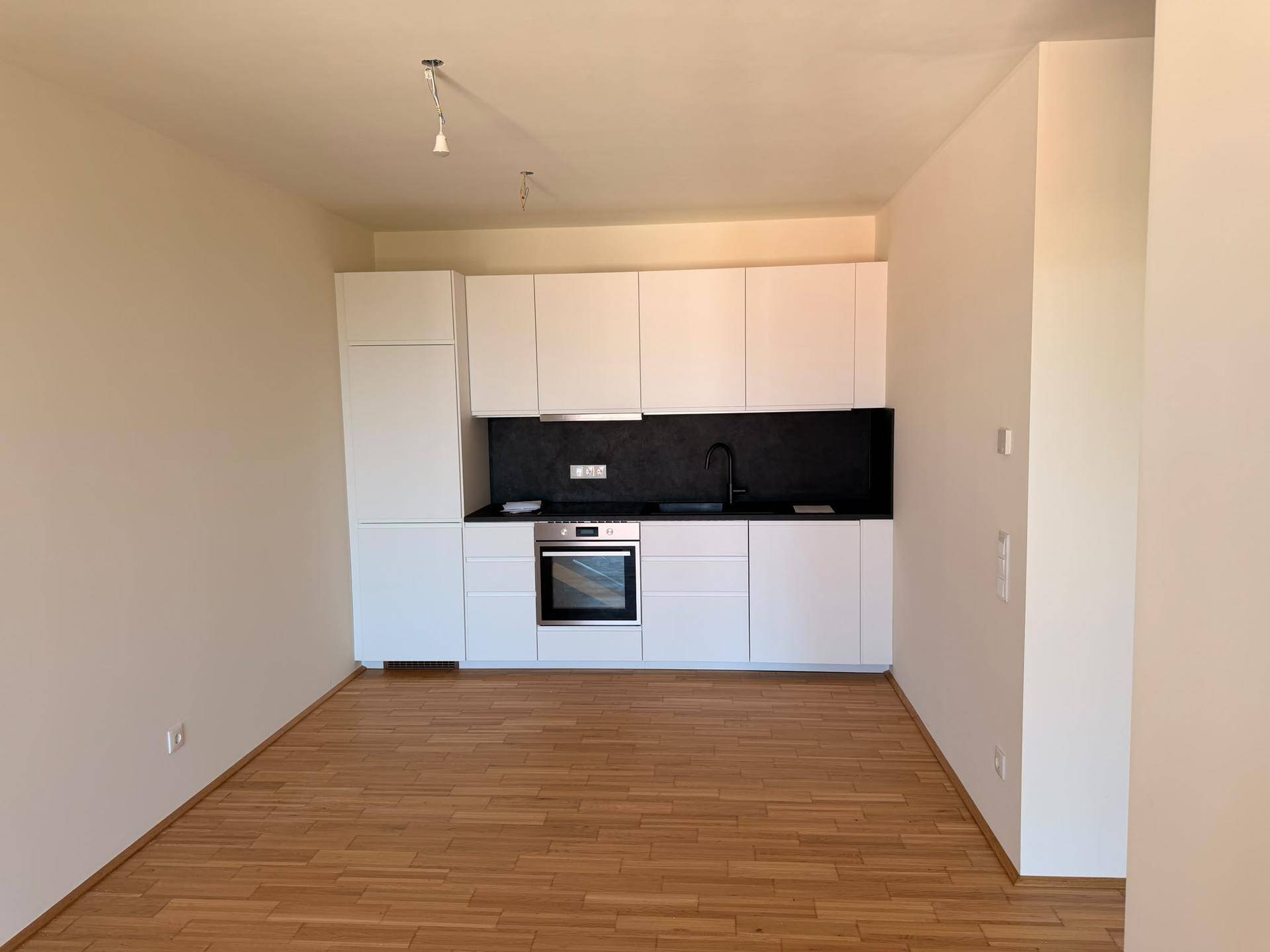 Moderne, leere Küche mit weißen Schränken, schwarzer Arbeitsplatte und Holzboden in einem offenen Raum.