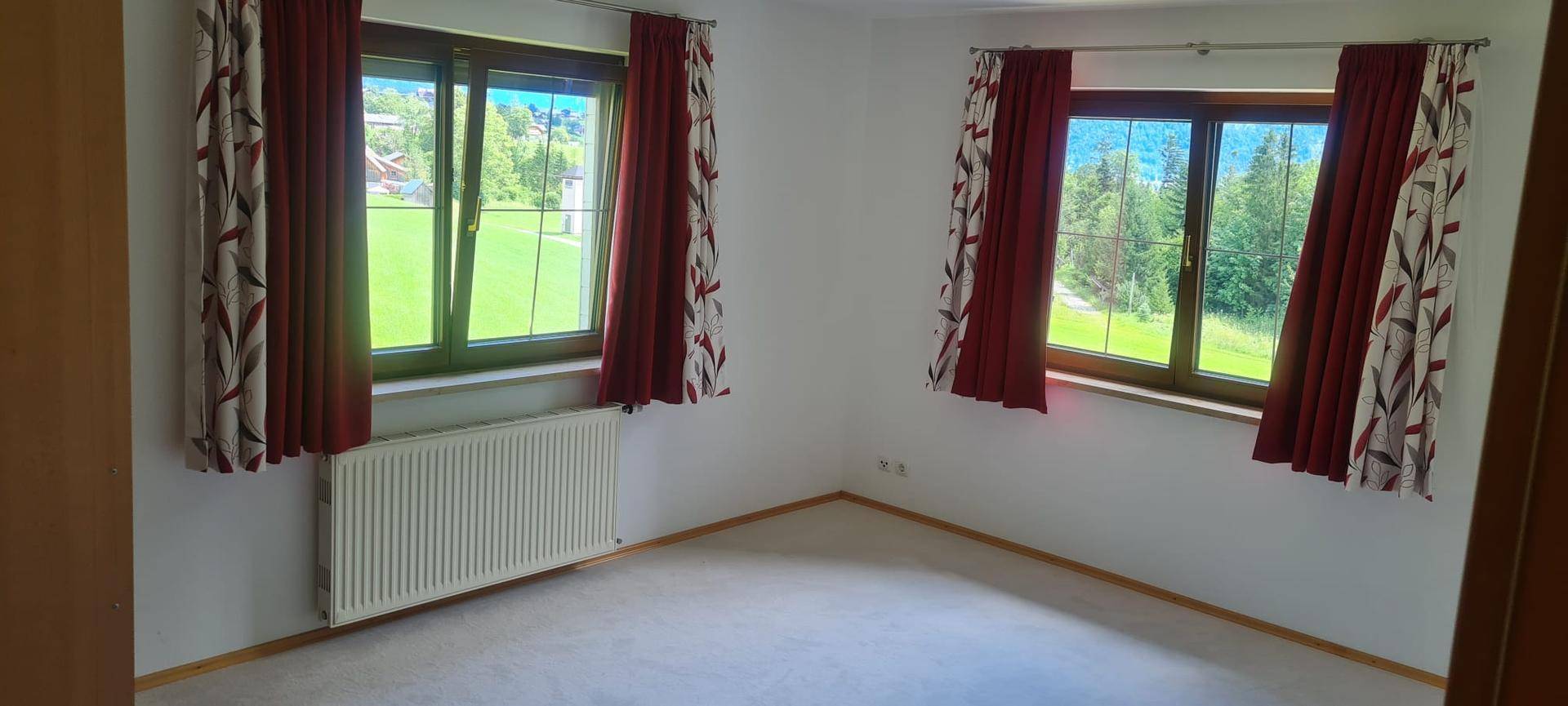Helles Zimmer mit zwei Fenstern, die einen grünen Ausblick bieten, und einem Heizkörper.
