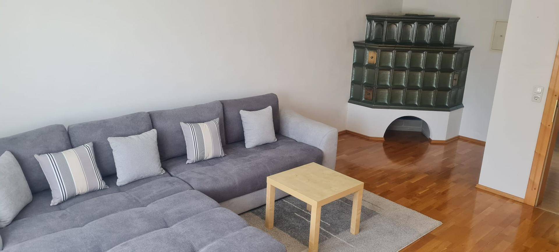 Geräumiges Wohnzimmer mit einem bequemen grauen Ecksofa und einem traditionellen Kachelofen.