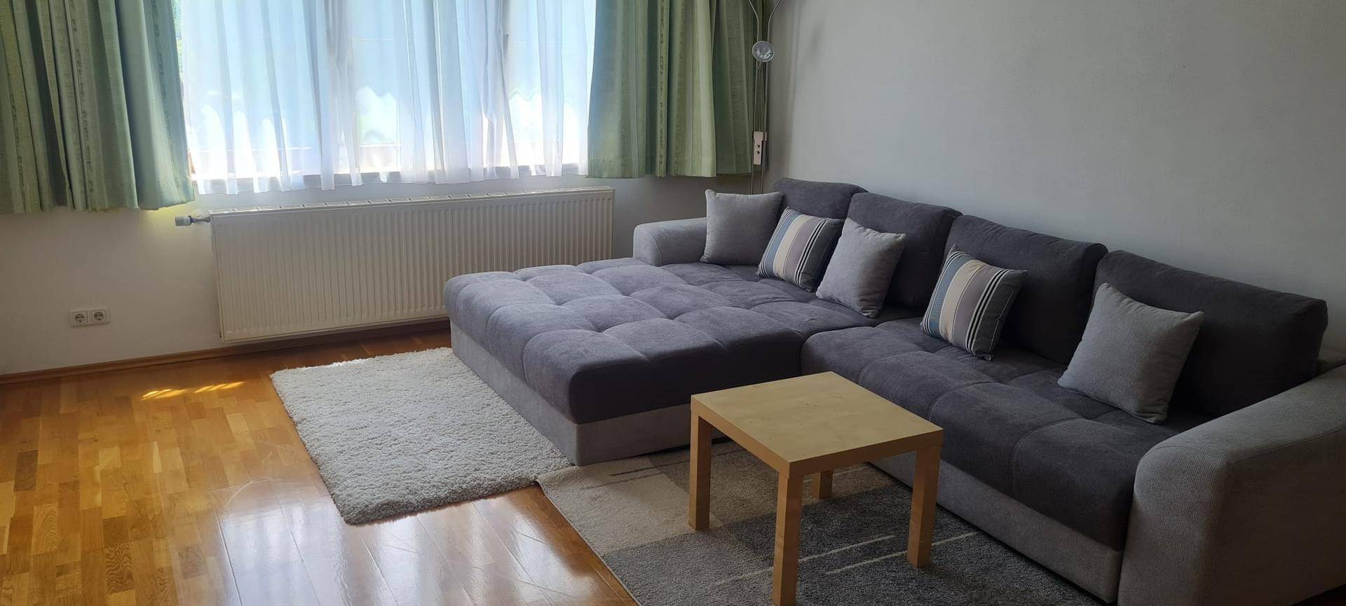 Modernes Wohnzimmer mit einem grauen Ecksofa, Holzfußboden und großen Fenstern.