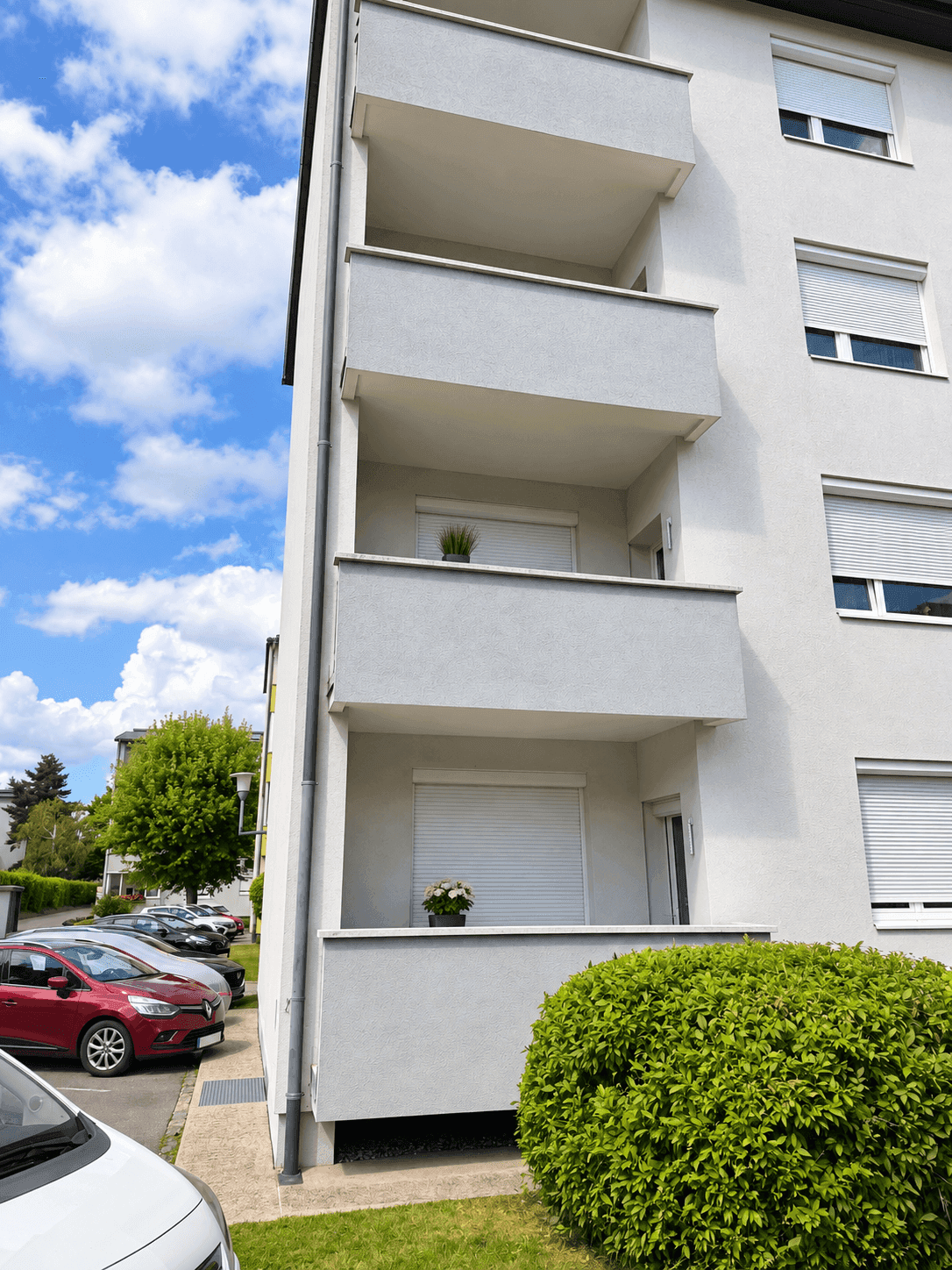 Moderne Wohnhausfassade mit mehreren Balkonen und Parkplätzen, umgeben von Grünflächen.