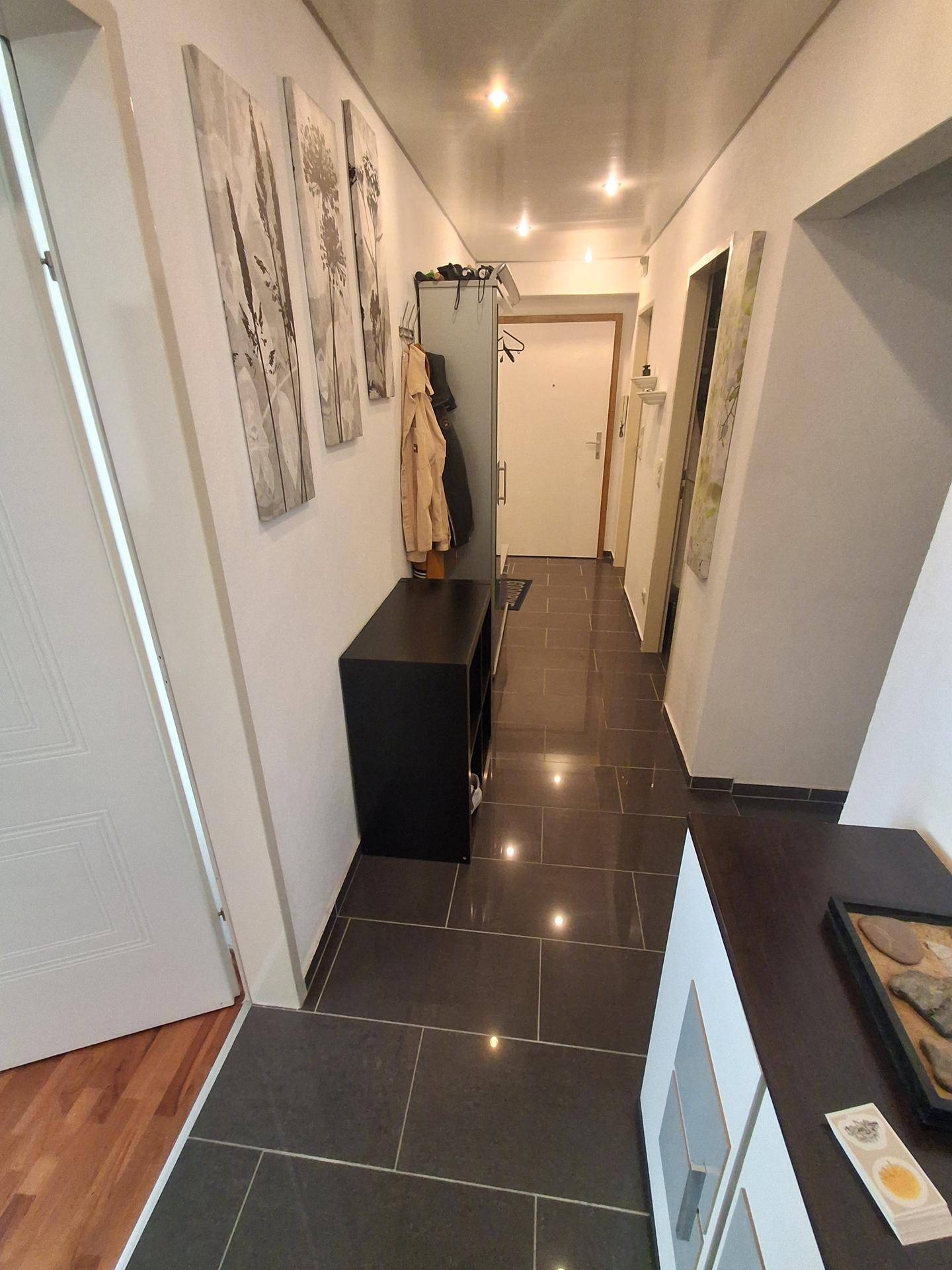 Langer Flur mit dunklen Fliesen, Garderobe und heller Beleuchtung.