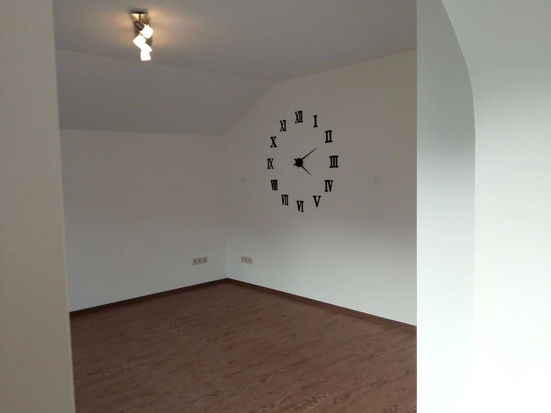 Leeres Zimmer mit Laminatboden, weißen Wänden und dekorativer Wanduhr unter einer Dachschräge.