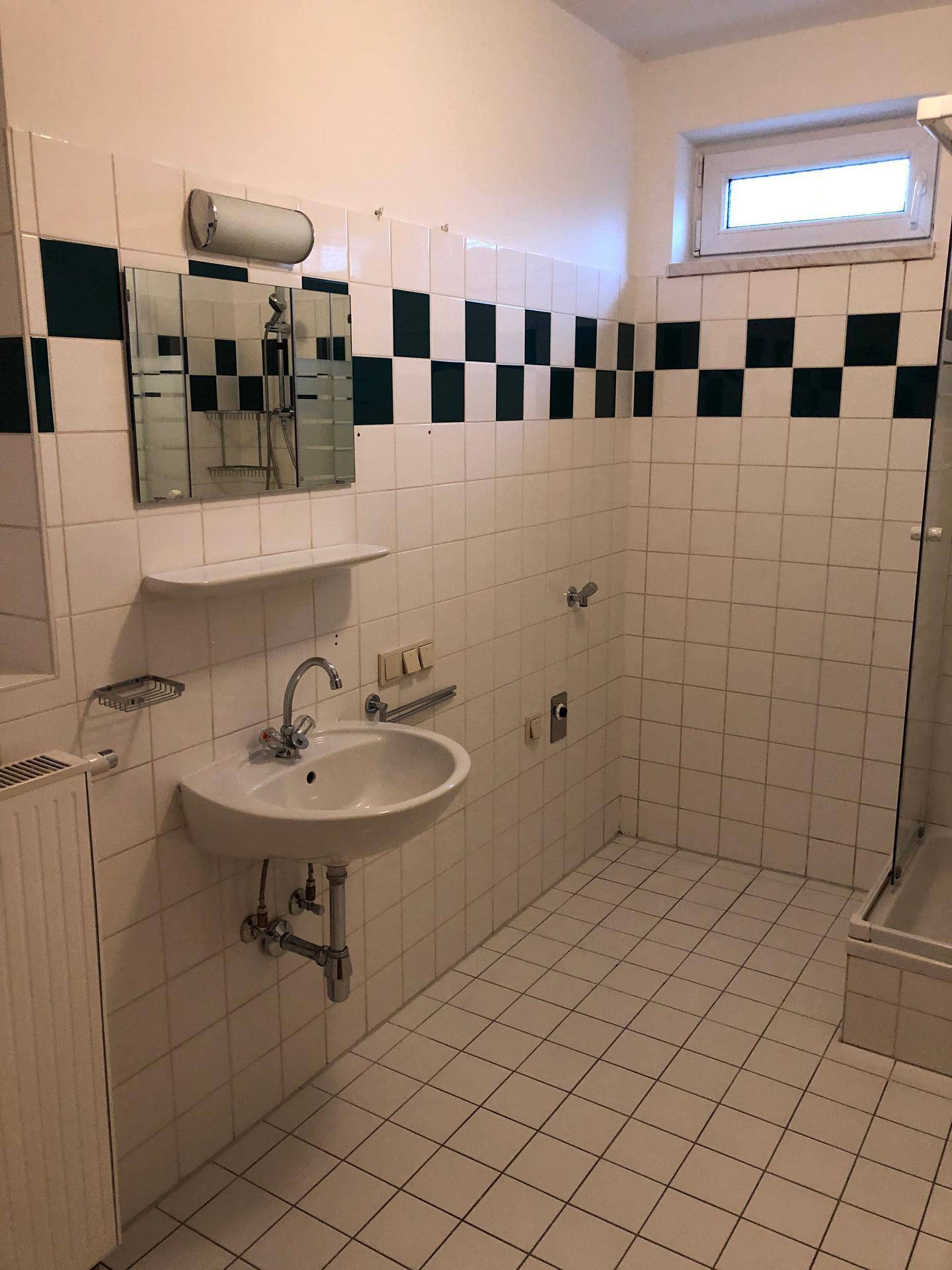 Badezimmer mit weiß-grünen Fliesen, Waschbecken, Spiegel und Heizkörper, ausgestattet mit einer Duschkabine.