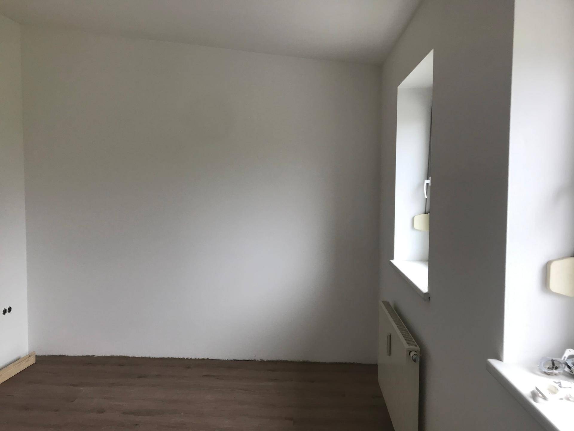 Leerer Raum mit weiß gestrichenen Wänden, Laminatboden und einem Fenster mit Heizkörper darunter.