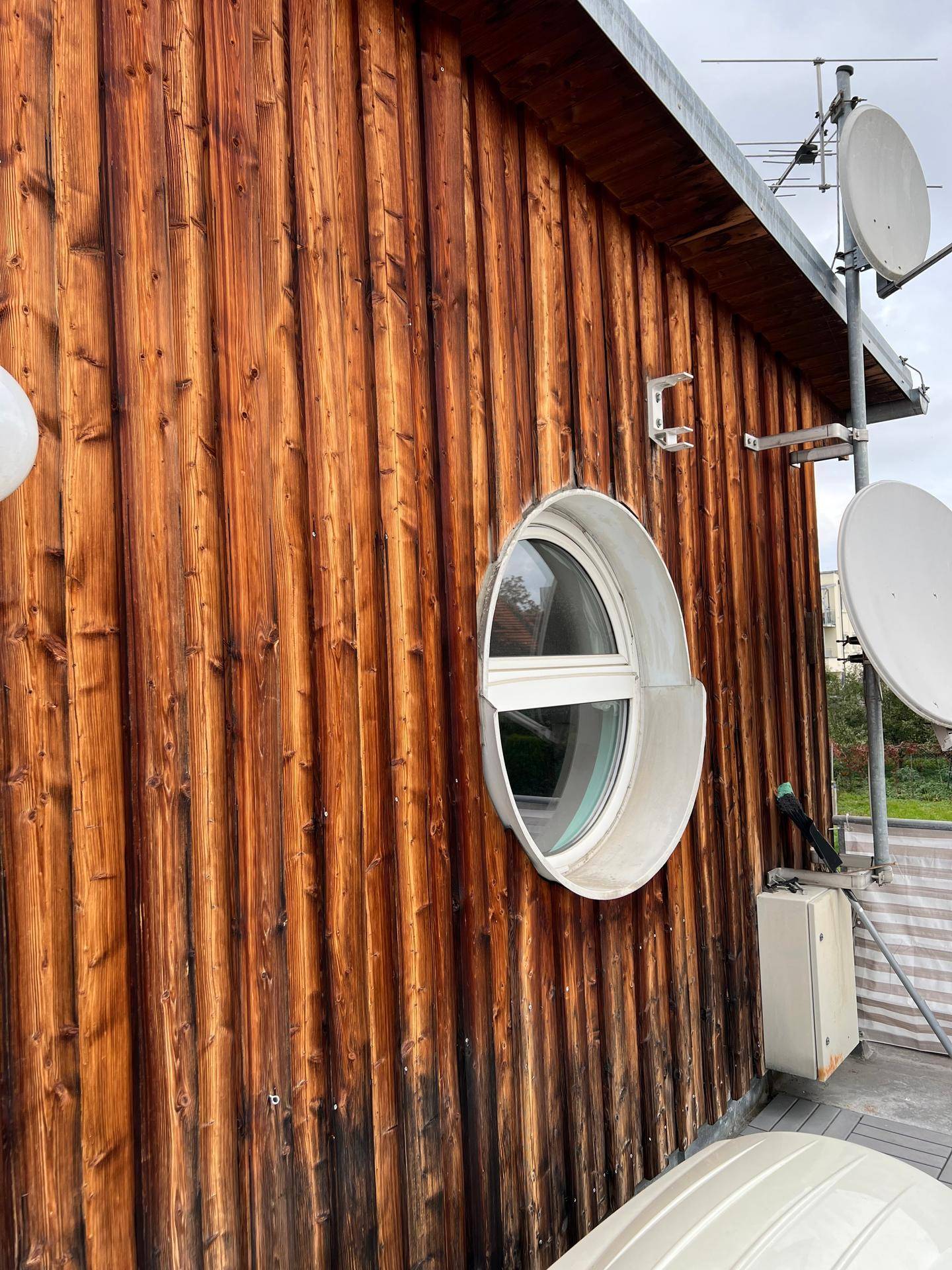 Holzverkleidete Fassade mit einem runden Fenster und mehreren Satellitenschüsseln auf dem Dach.