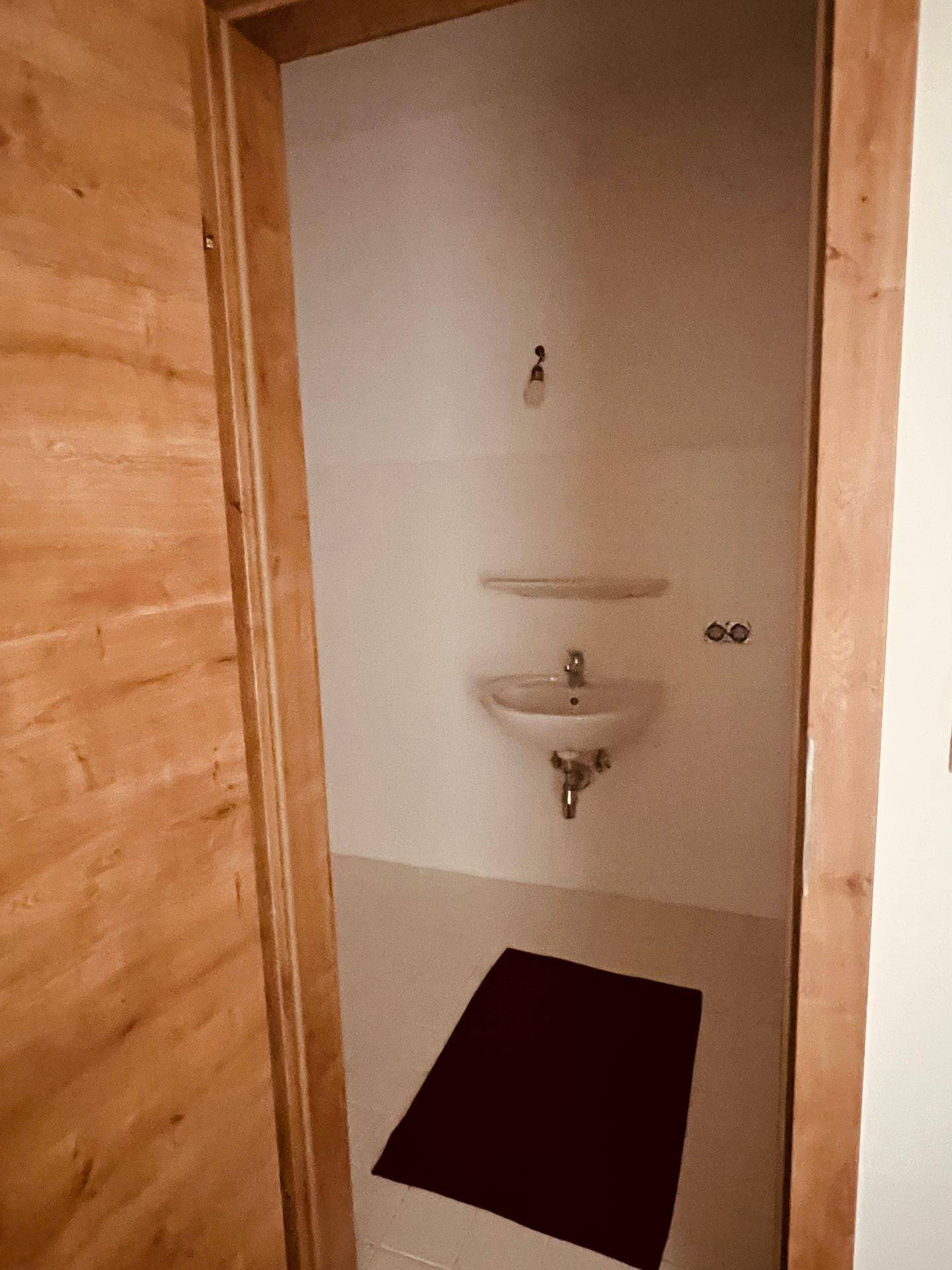 Kleines Gäste-WC mit Waschbecken und schlichten weißen Fliesen, durch eine Holztür zugänglich.