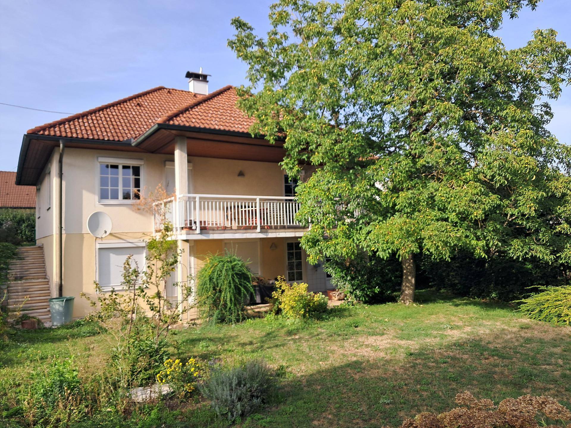 Seitenansicht des Hauses mit Balkon, umgeben von einem großen Baum und viel Grün.