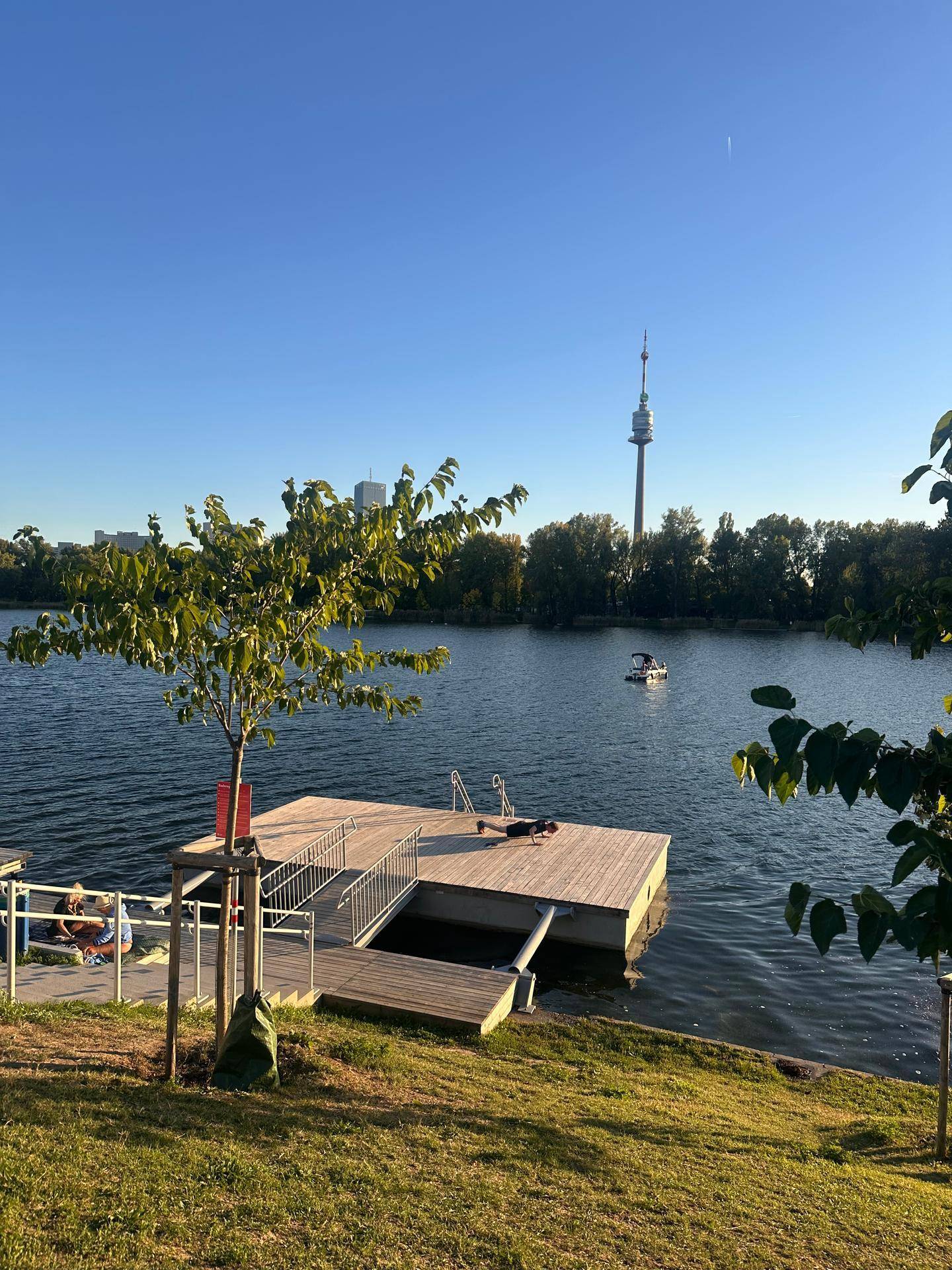 Ruhiger Seeblick mit Holzsteg, Boot und dem Donauturm sowie der Skyline in der Ferne.