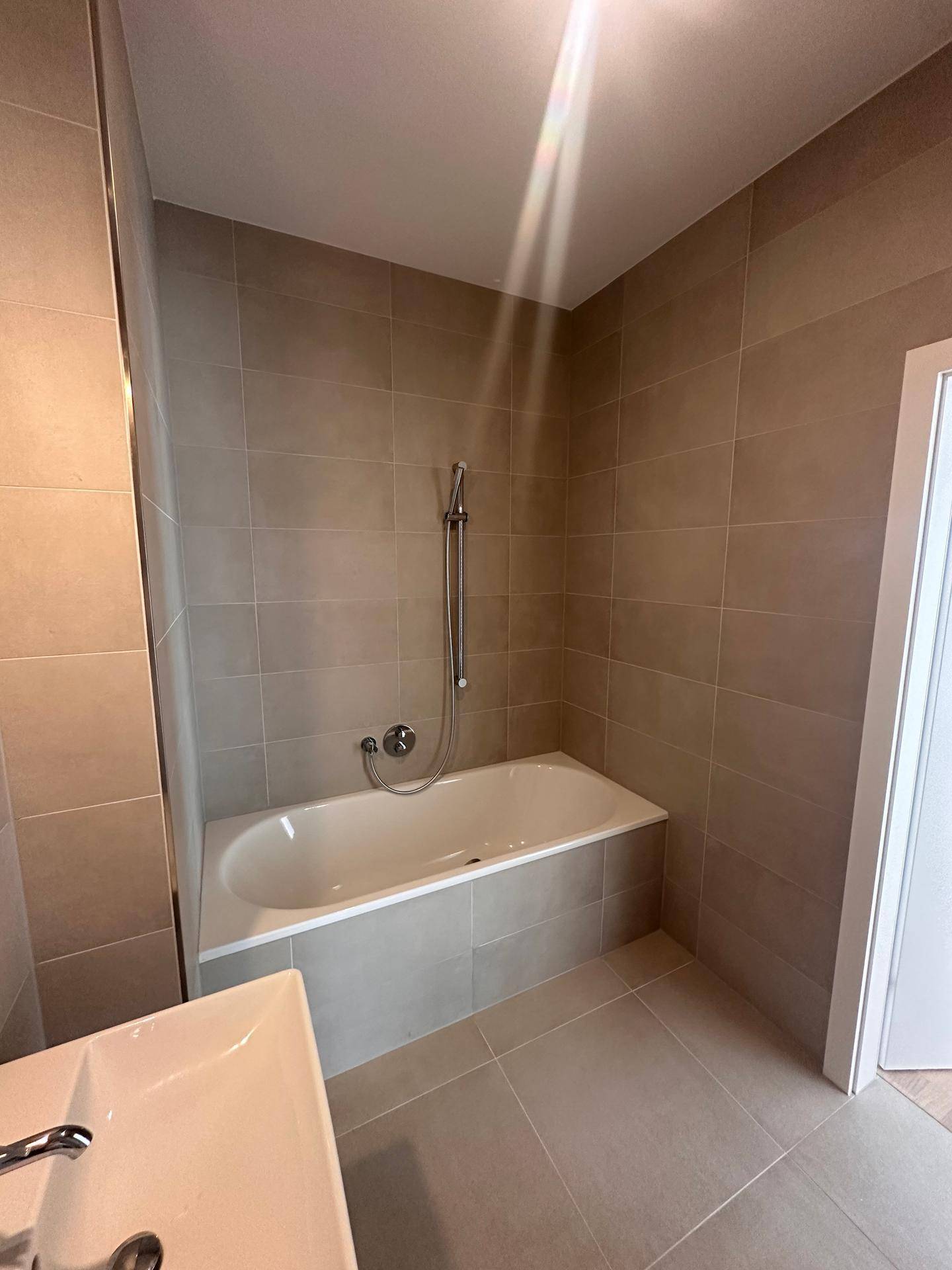 Badezimmer mit eingebauter Badewanne und Duschfunktion, umgeben von modernen hellgrauen Fliesen.