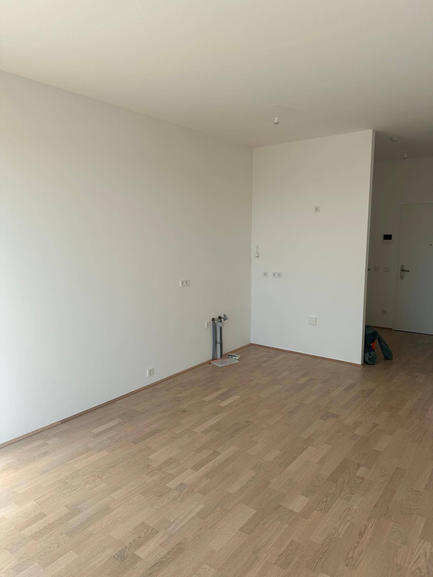 Leerer Küchenbereich mit Anschlüssen für eine Einbauküche und hellem Parkettboden.