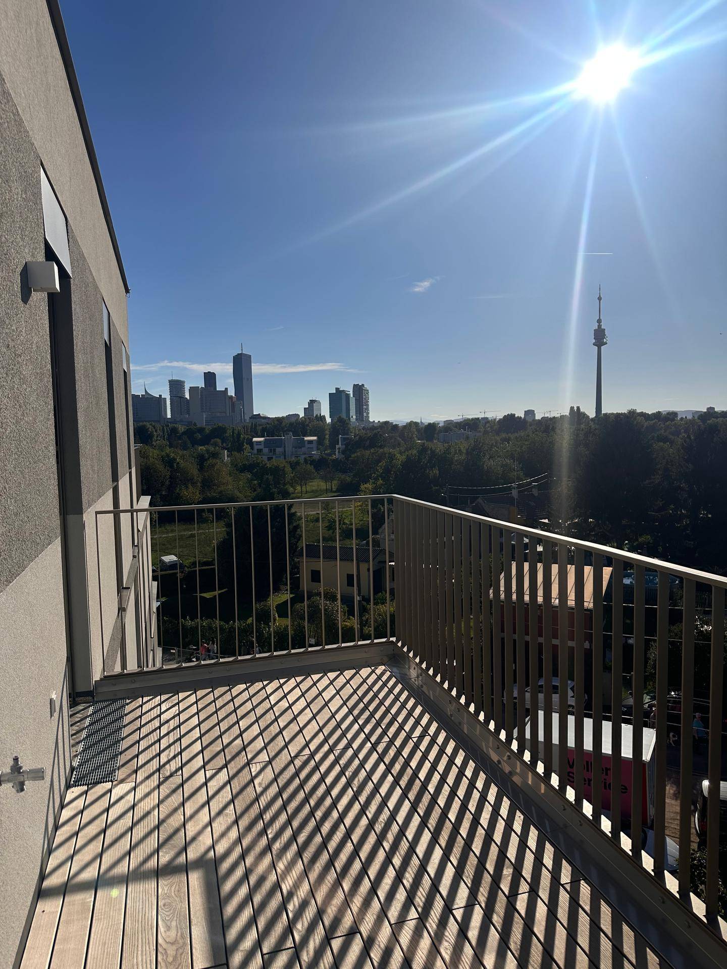 Sonniger Balkon mit Holzboden und Blick auf die Wiener Skyline und den Donauturm.