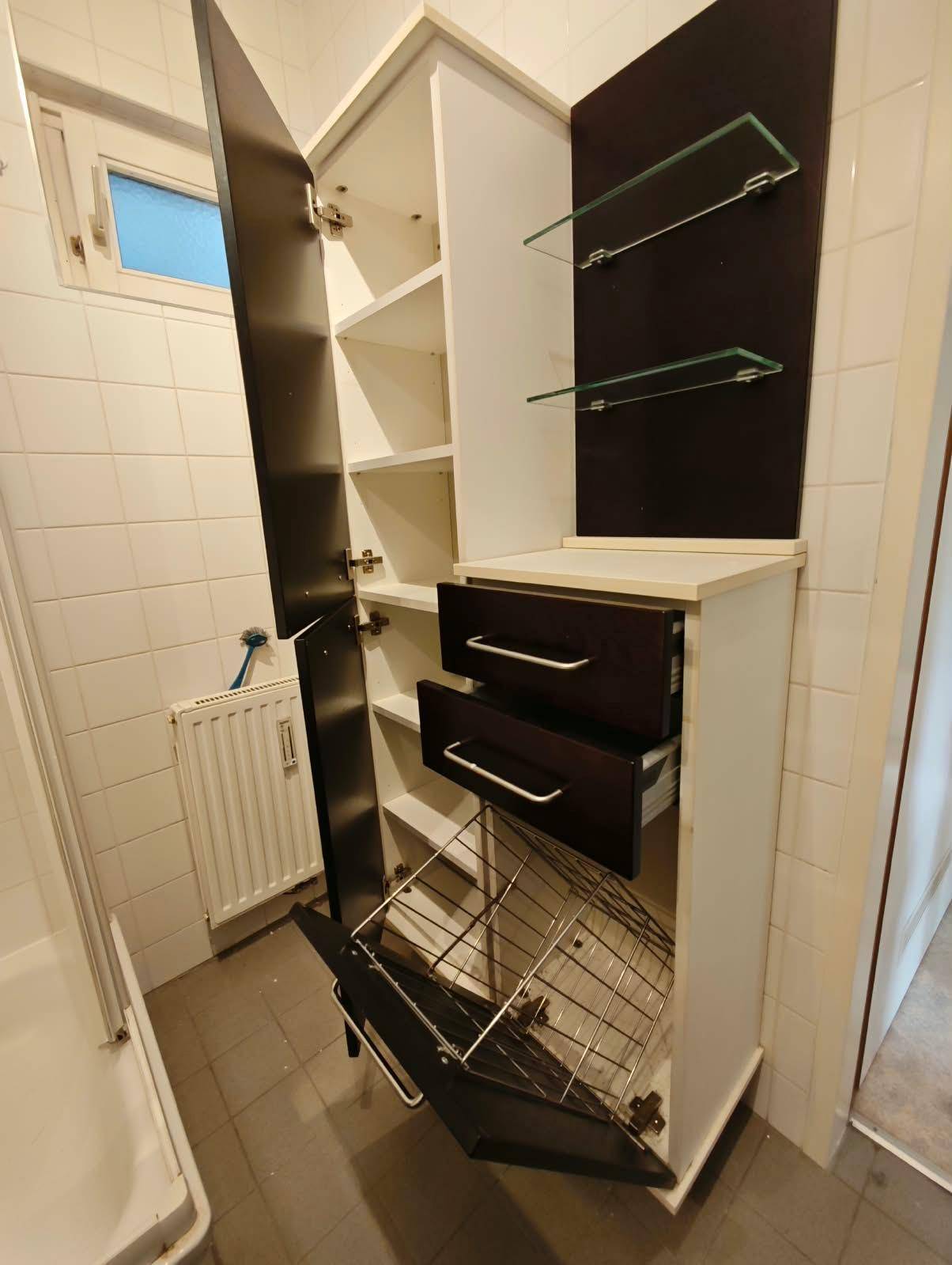 Badezimmerschrank mit offenen Fächern und integriertem Wäschekorb, neben einem Fenster.