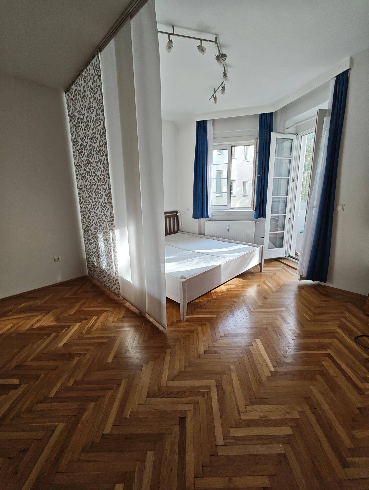 Schlafzimmer mit Fischgrätparkett, Bett und direktem Zugang zur verglasten Loggia.