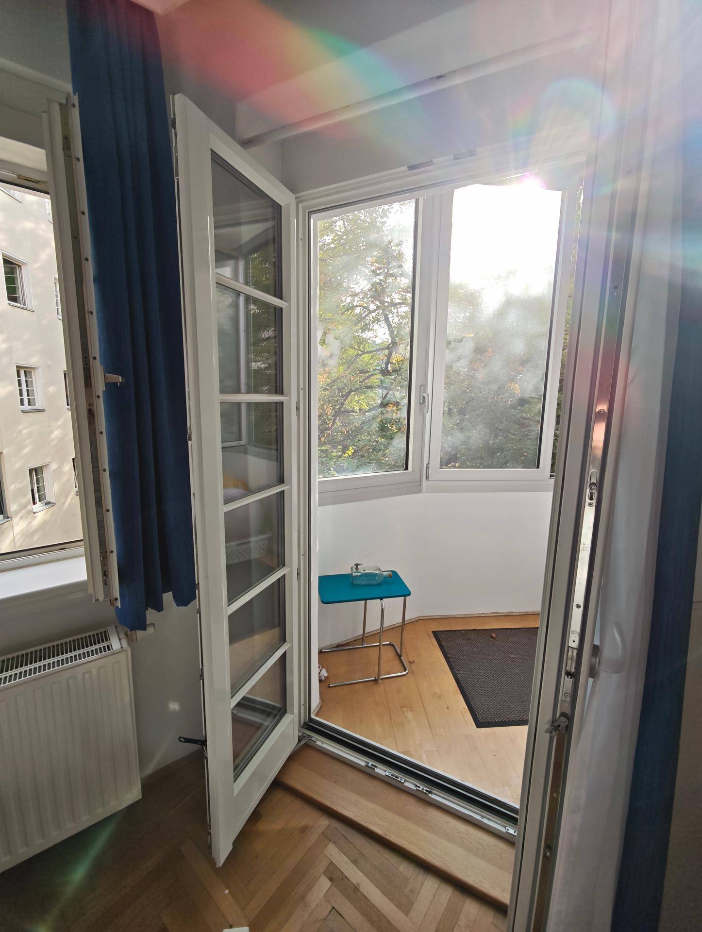 Heller Raum mit Holzboden und Zugang zu einem kleinen Balkon mit Blick ins Grüne.