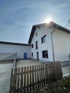 Seitenansicht des weißen Wohnhauses mit Balkon und angrenzendem Nebengebäude unter blauem Himmel.