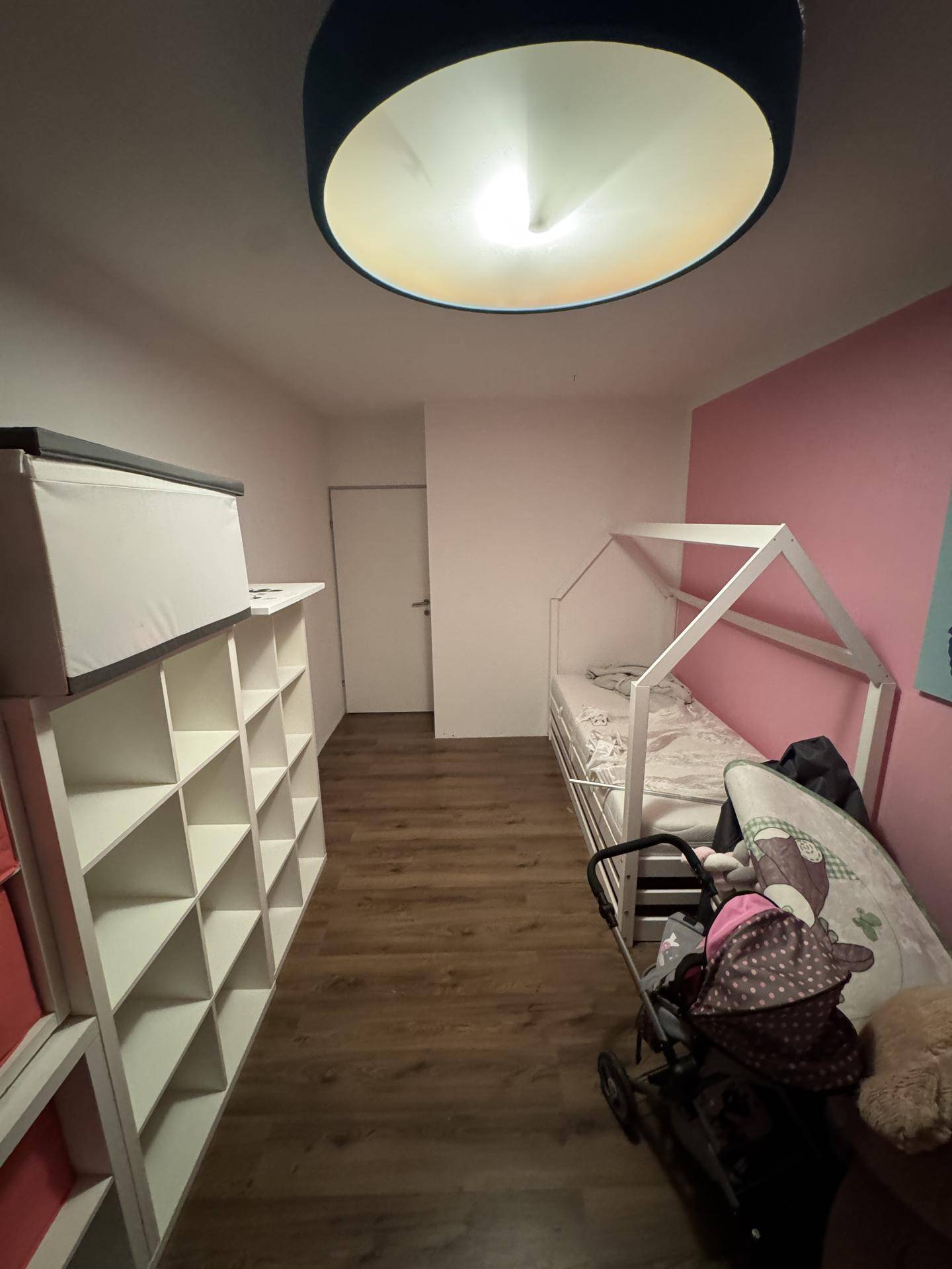 Kinderzimmer mit Laminatboden, rosa Wand und einem Regal für Spielzeug oder Bücher, gut organisiert.