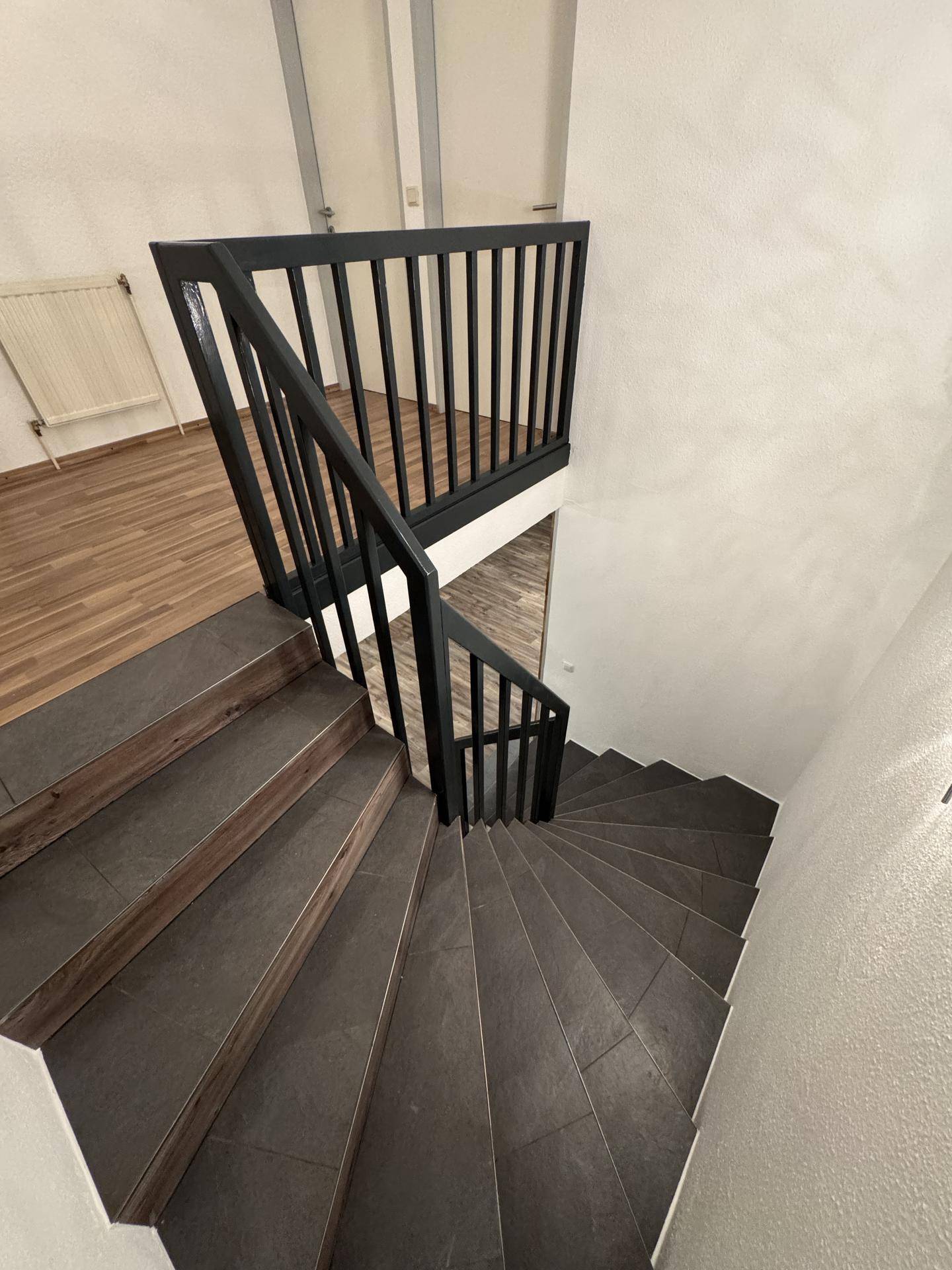 Wendeltreppe mit dunklen Fliesenstufen und schwarzem Geländer, die in die obere Etage führt.