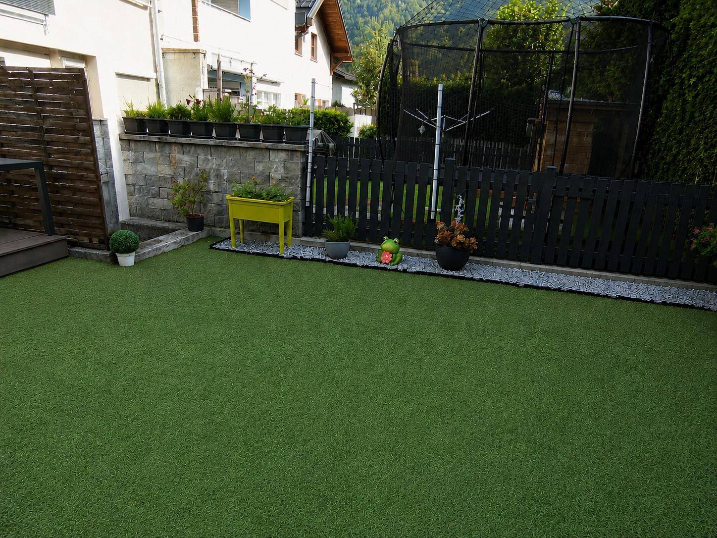 Gepflegter Gartenbereich mit Kunstrasen, kleiner Holzterrasse und Trampolin im Hintergrund.