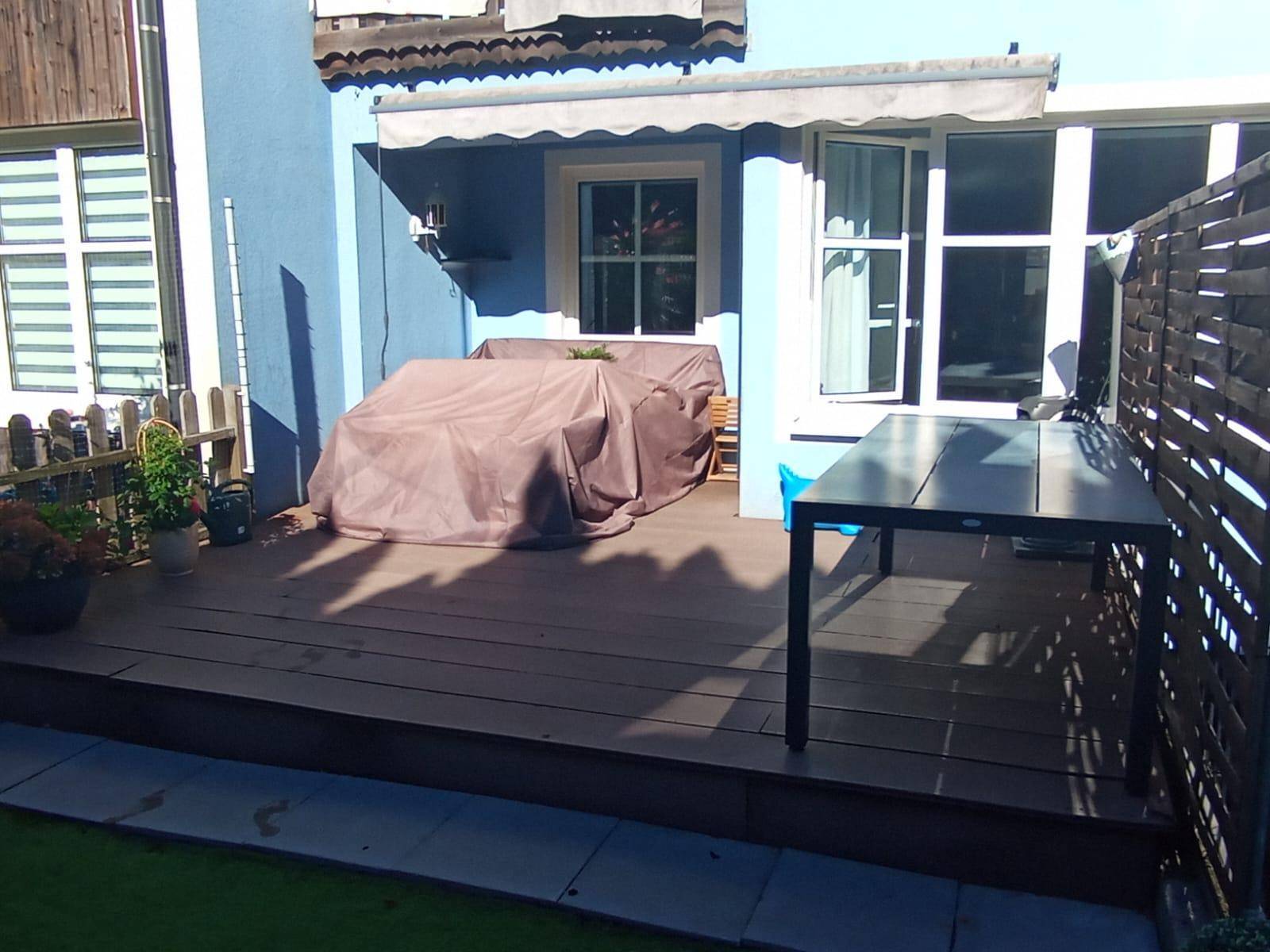 Geräumige Holzterrasse mit Esstisch, abgedeckten Gartenmöbeln und blauer Hausfassade.