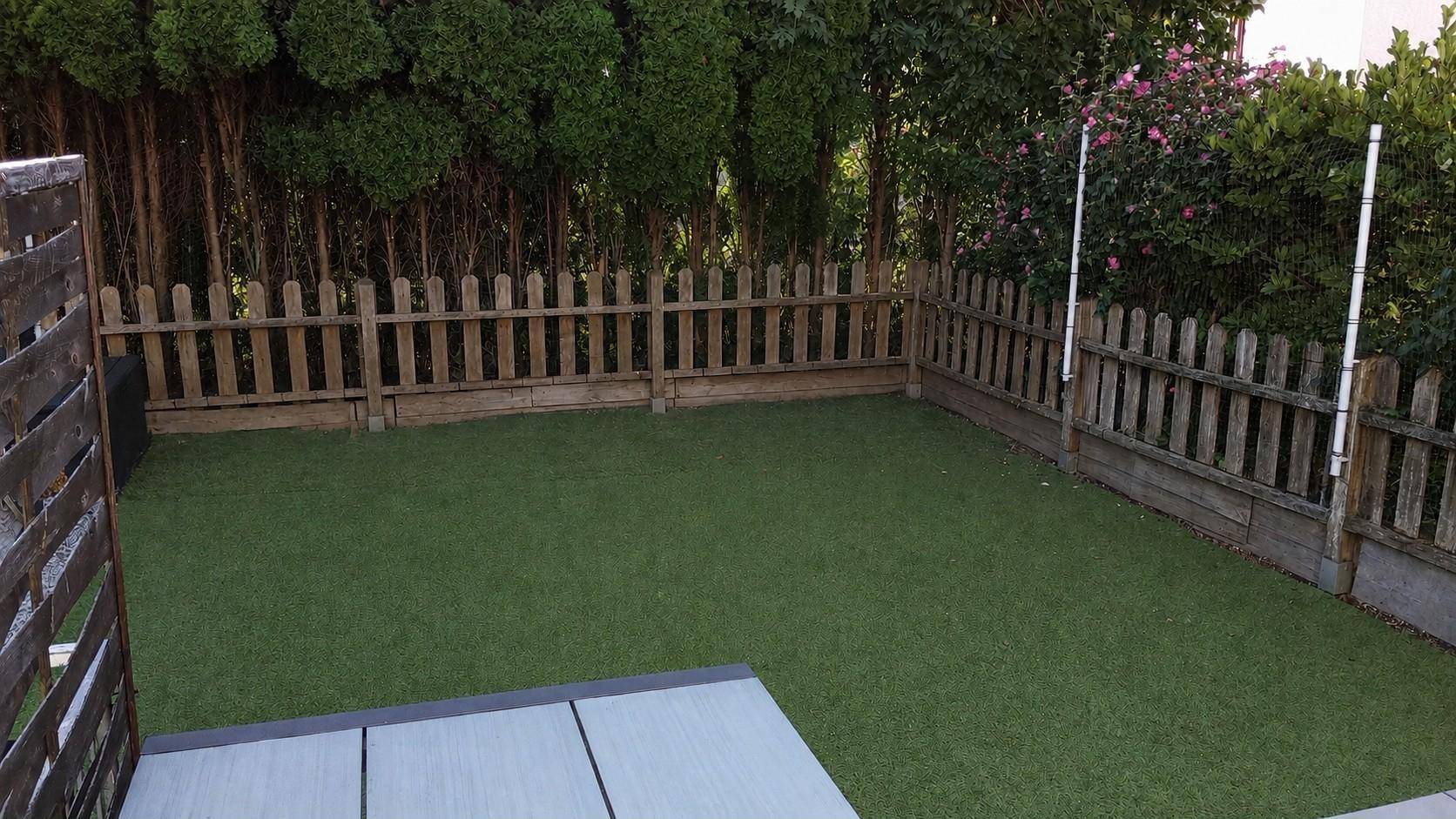 Gepflegter Gartenbereich mit Kunstrasen und Holzzaun, umgeben von Bäumen und Sträuchern.
