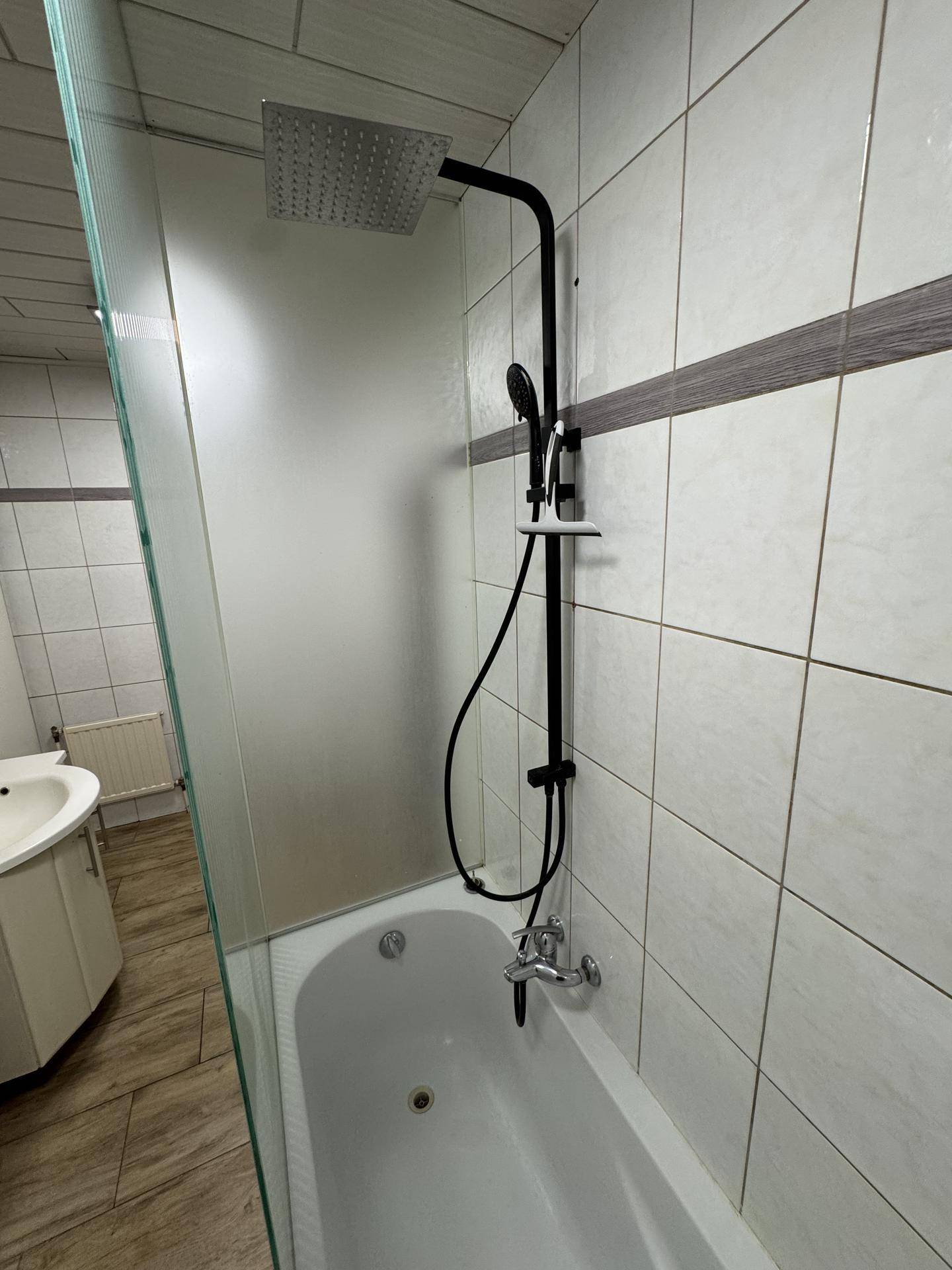Duschbadewanne mit moderner Regendusche und Fliesenwand, abgetrennt durch eine Glaswand.