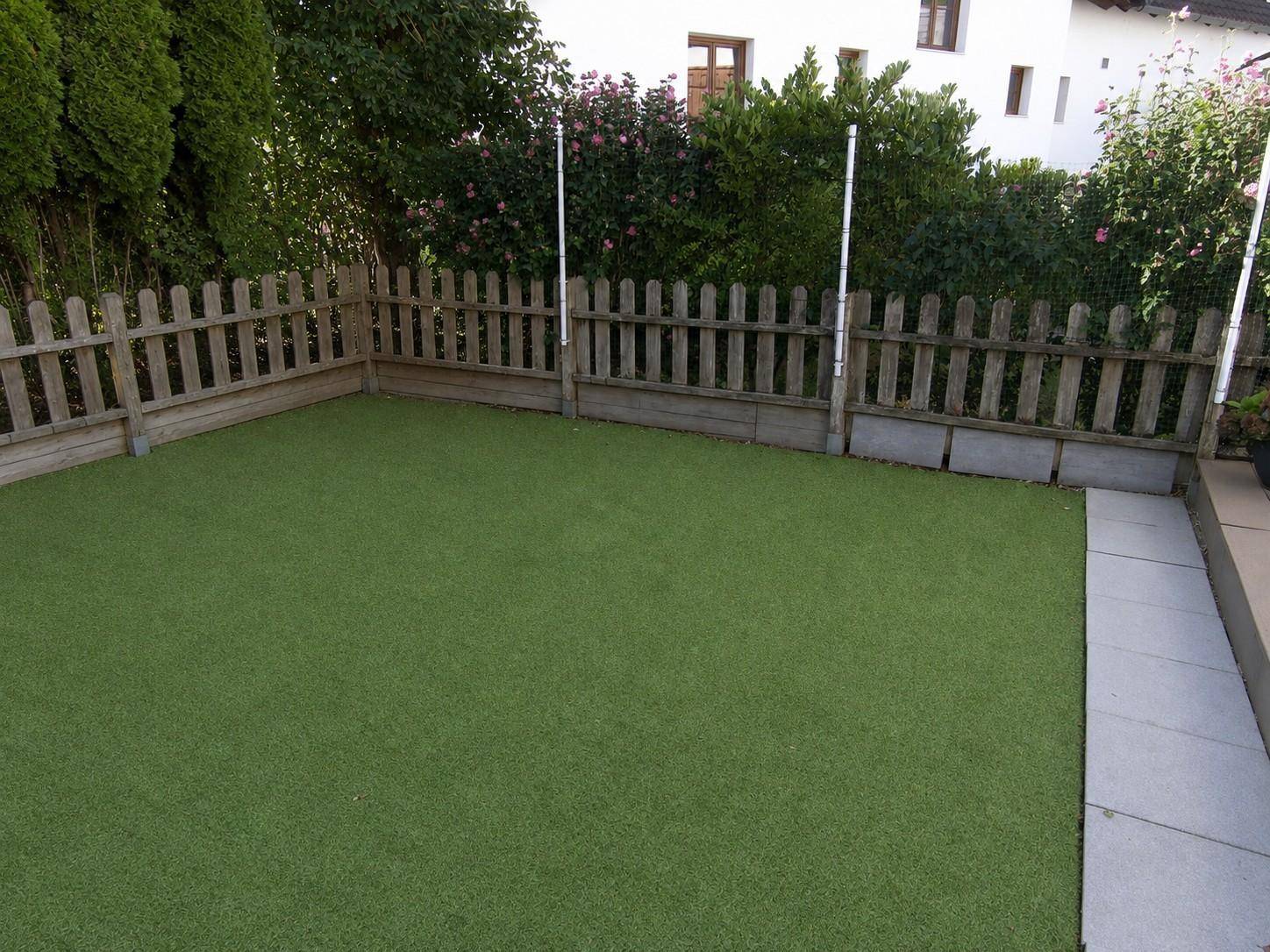 Kleiner, eingezäunter Garten mit Kunstrasen und angrenzendem Wohnhaus, ideal für Entspannung.