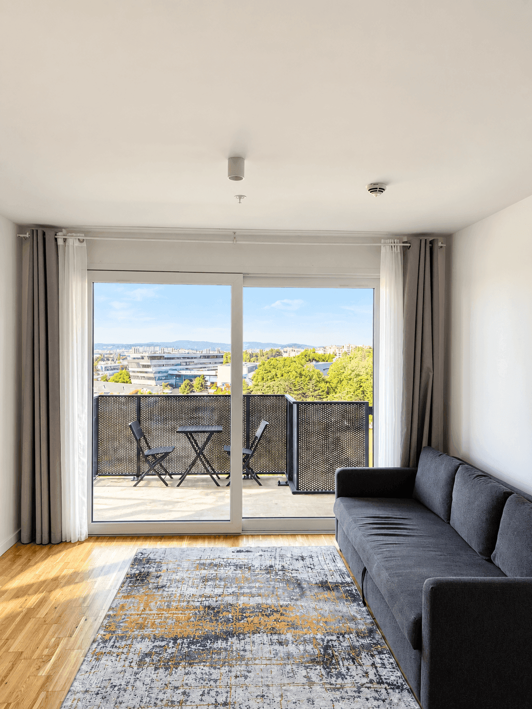 Helles Wohnzimmer mit grauem Sofa, Teppich und Balkonzugang mit Stadtblick.
