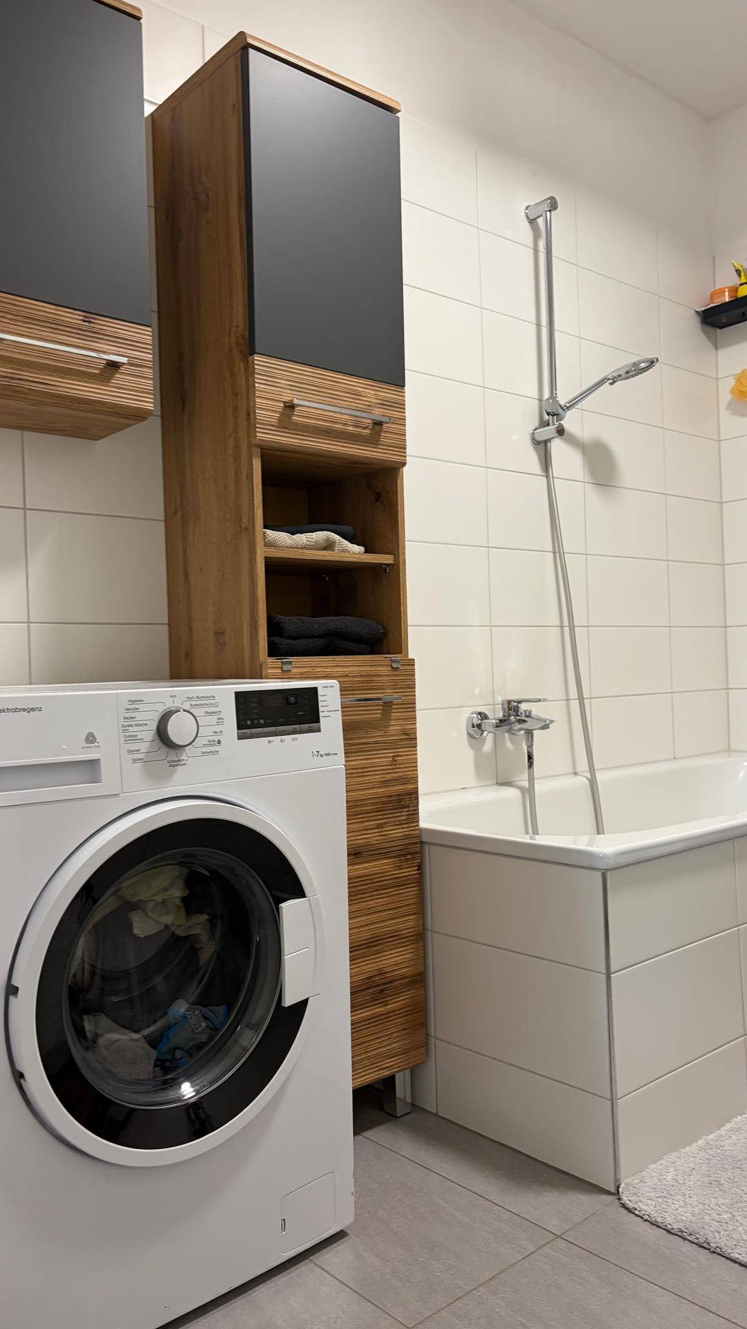 Badewanne und Waschmaschine in einem hell gefliesten Badezimmer mit Holzschränken.