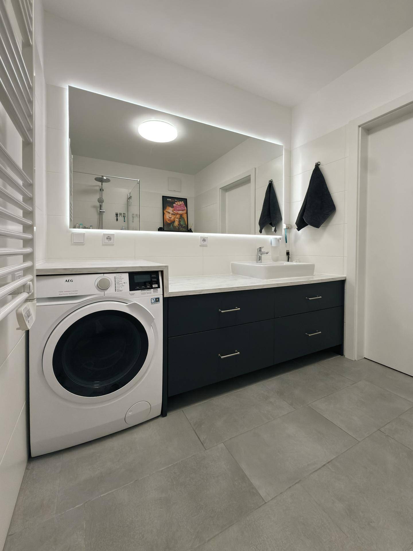 Modernes Badezimmer mit großem beleuchtetem Spiegel, Waschmaschine und dunklem Unterschrank.