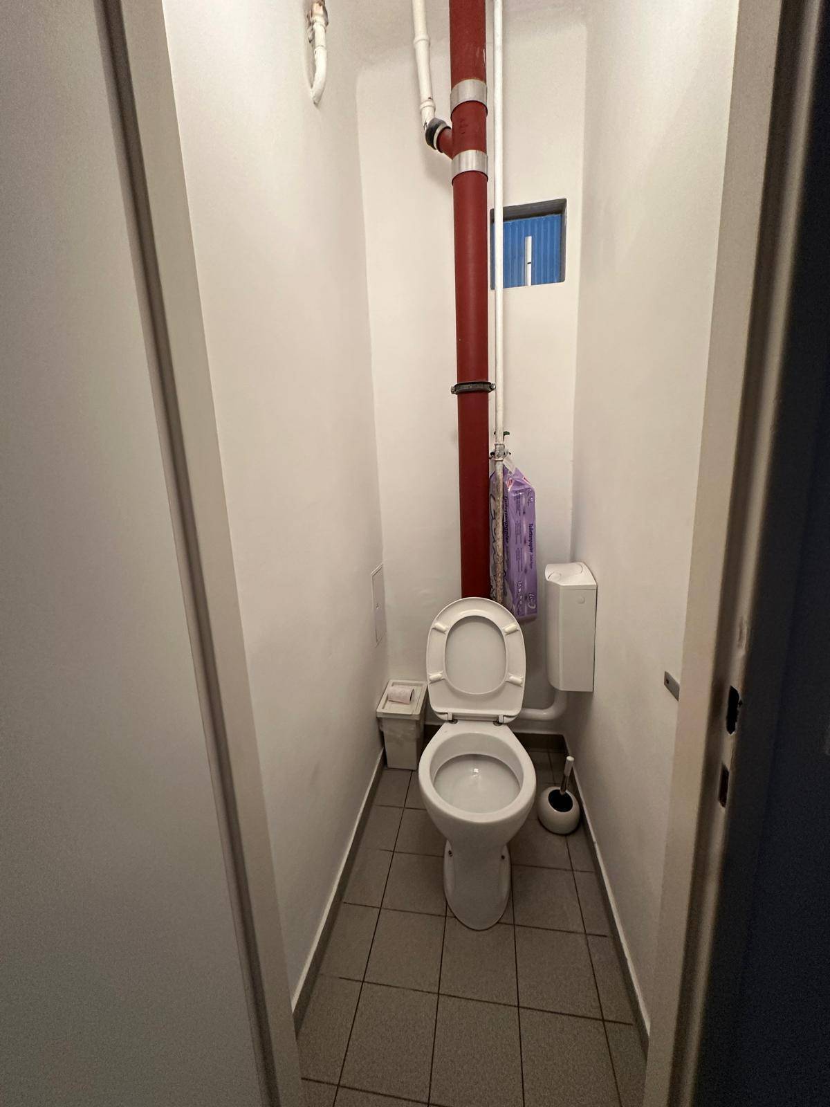 Kleines separates WC mit Fliesenboden und sichtbaren Rohrleitungen, einfach ausgestattet.