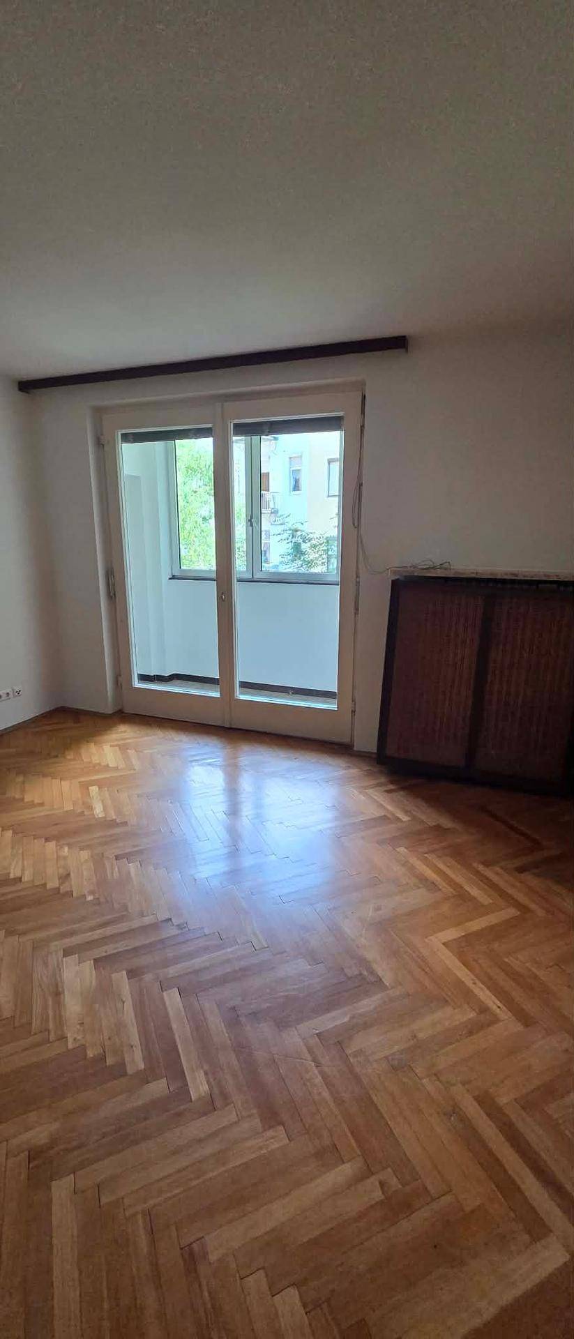Geräumiges Zimmer mit Fischgrätparkett und Zugang zu einem Balkon durch eine große Schiebetür.
