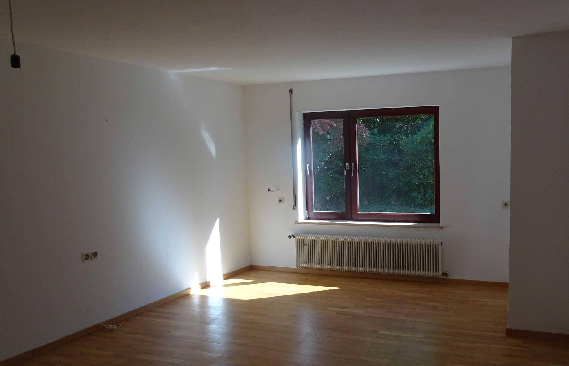 Helles, leeres Zimmer mit Holzboden und großem Fenster, das viel Sonnenlicht hereinlässt.