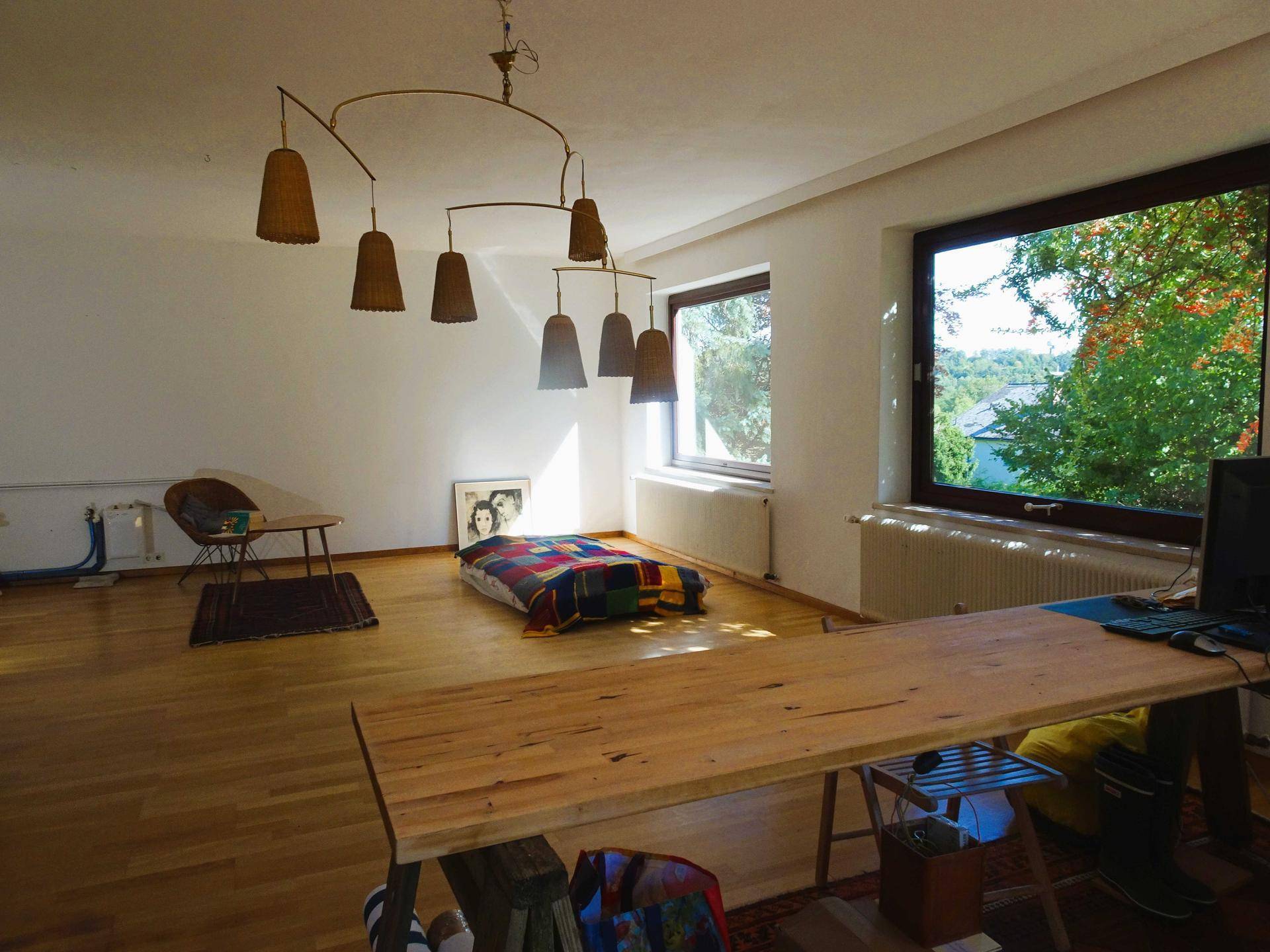 Geräumiges Wohnzimmer mit Holzboden, moderner Deckenleuchte und viel Tageslicht.