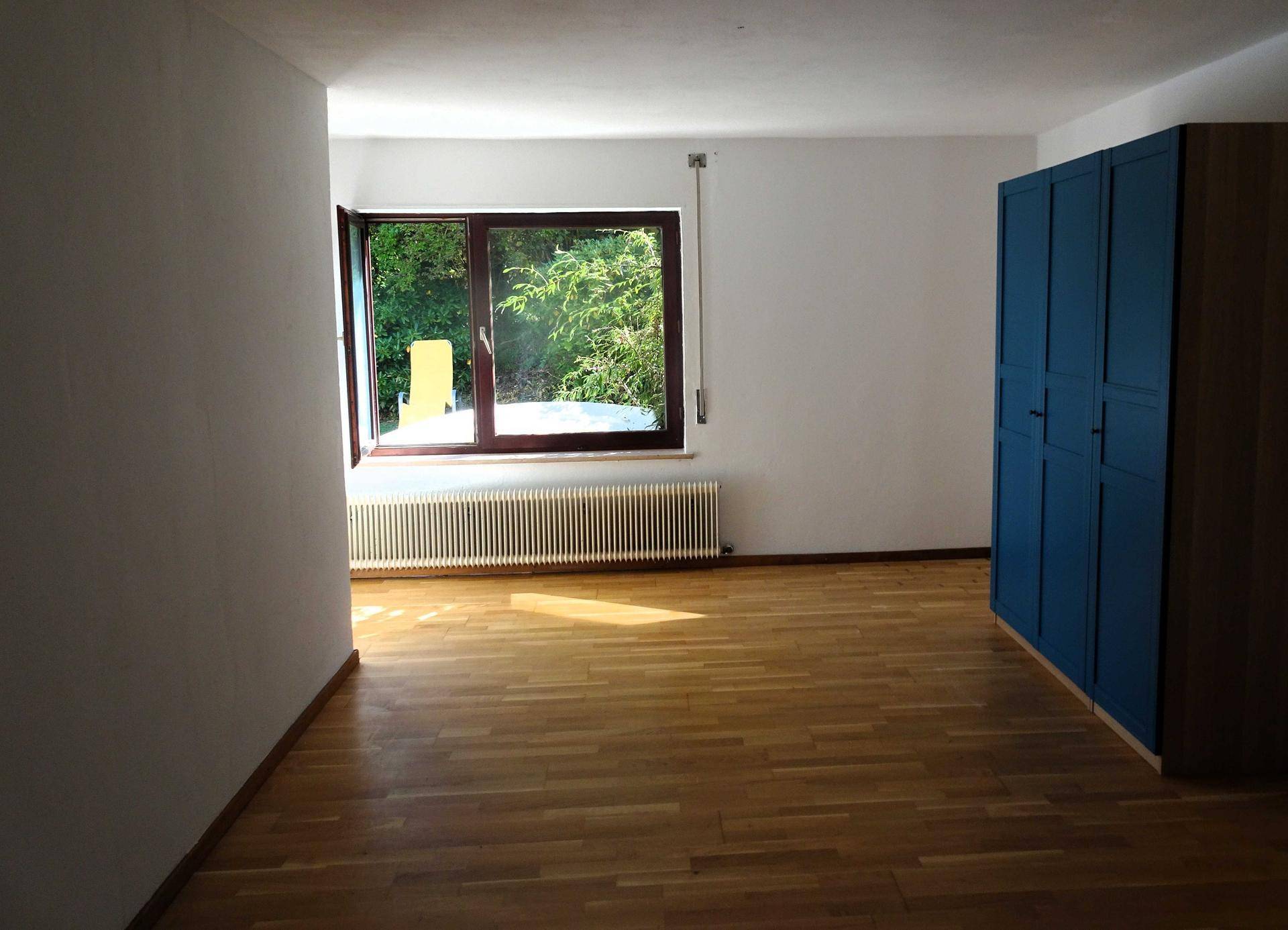 Leeres Schlafzimmer mit Holzboden, blauer Kommode und Fenster mit Blick ins Grüne.