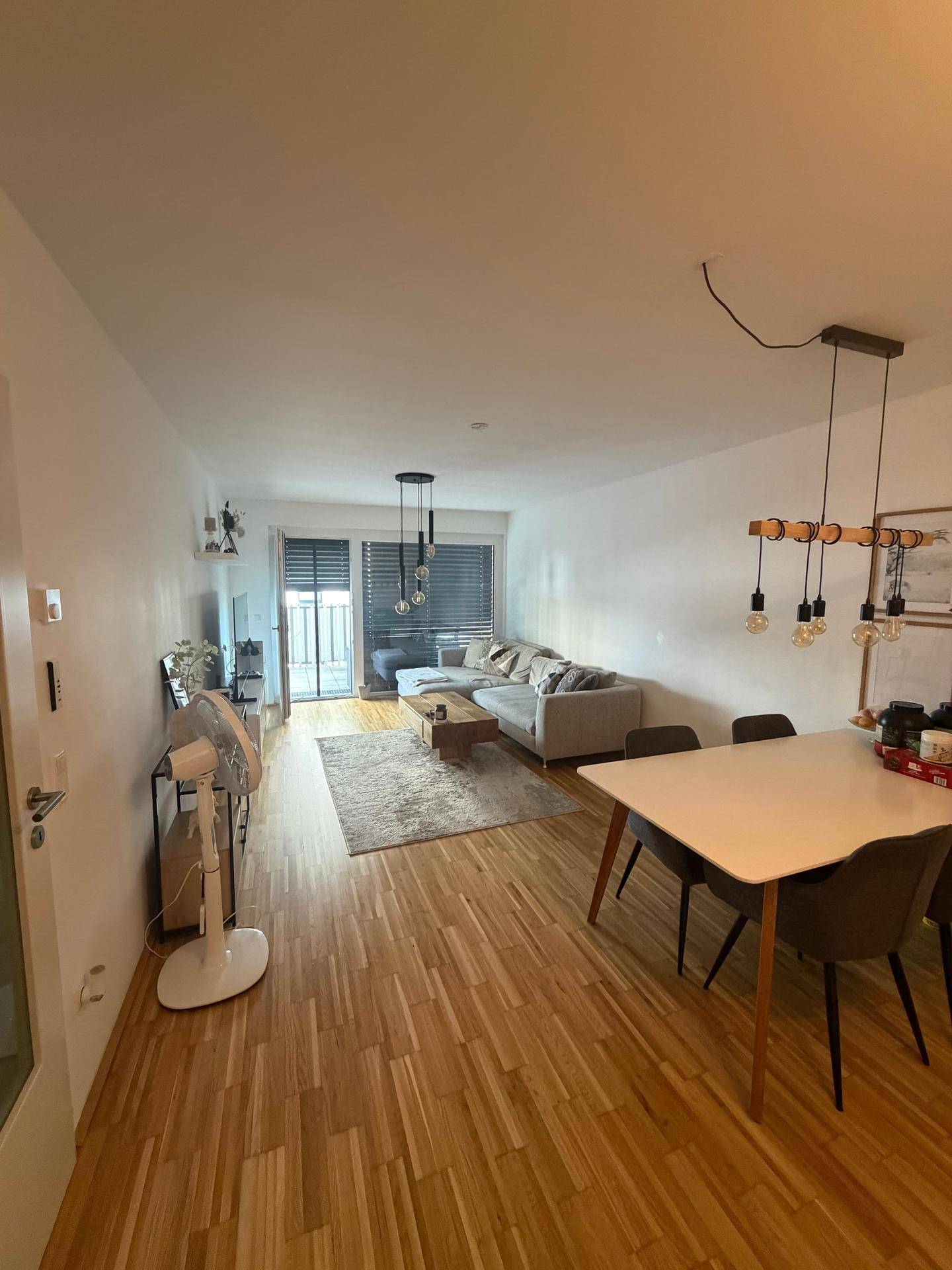 Geräumiger Wohn-Essbereich mit Holzparkett, großem Sofa und Esstisch, sowie Zugang zum Balkon.