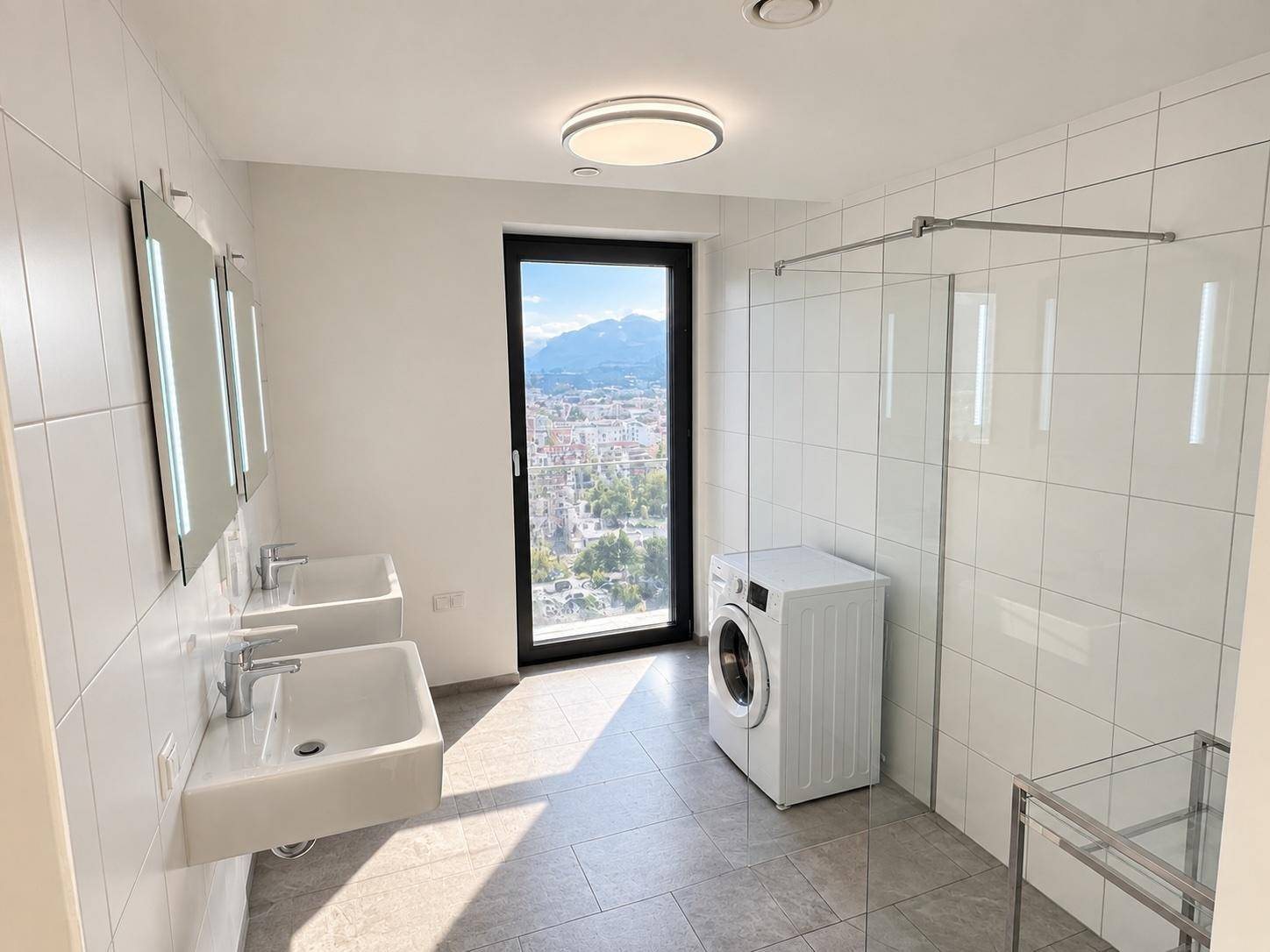 Modernes Badezimmer mit Doppelwaschbecken, Dusche, Waschmaschine und Bergblick durch das Fenster.