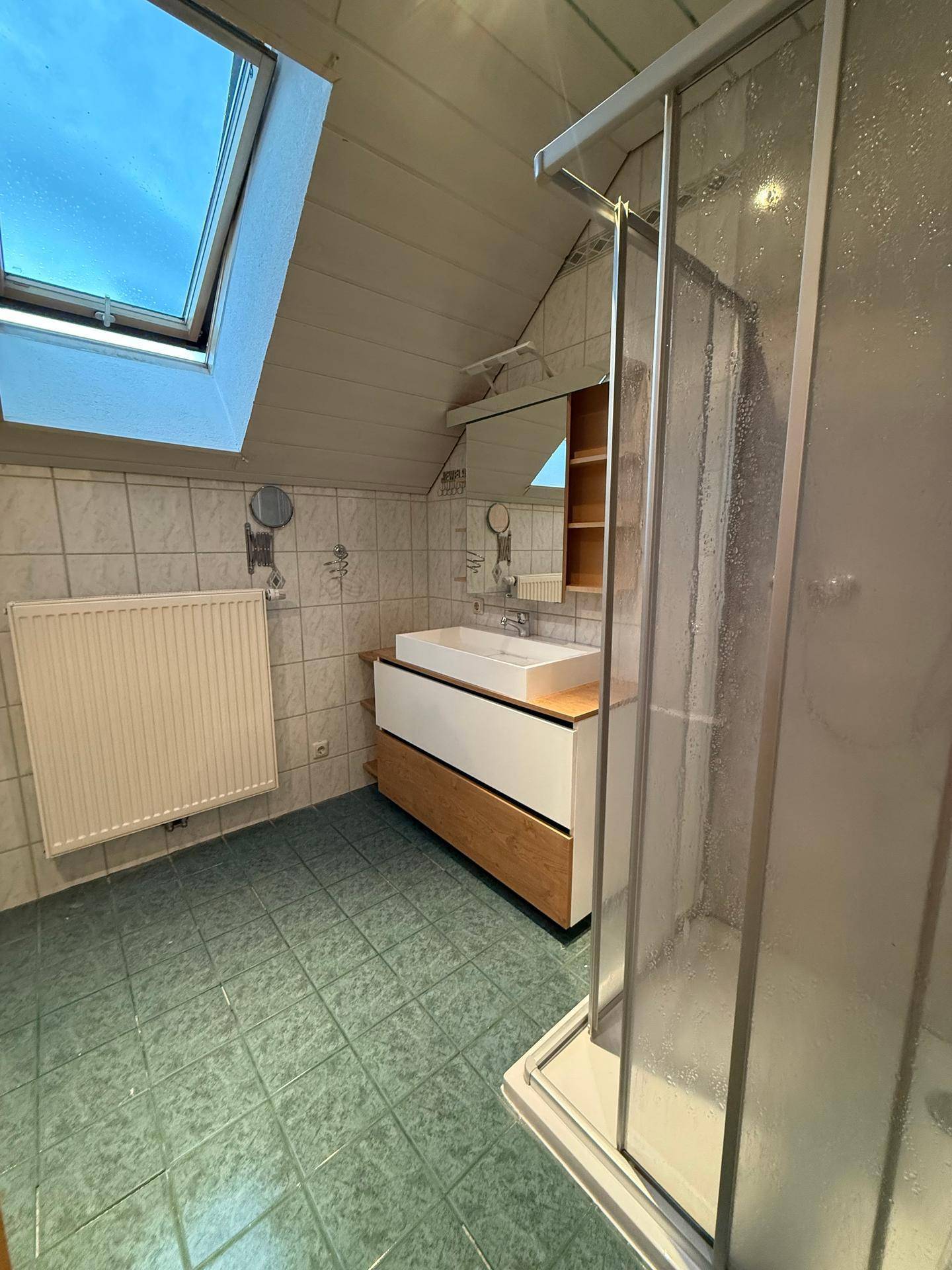 Badezimmer mit Dusche, Waschtisch, Dachfenster und grünlichen Fliesen unter der Dachschräge.