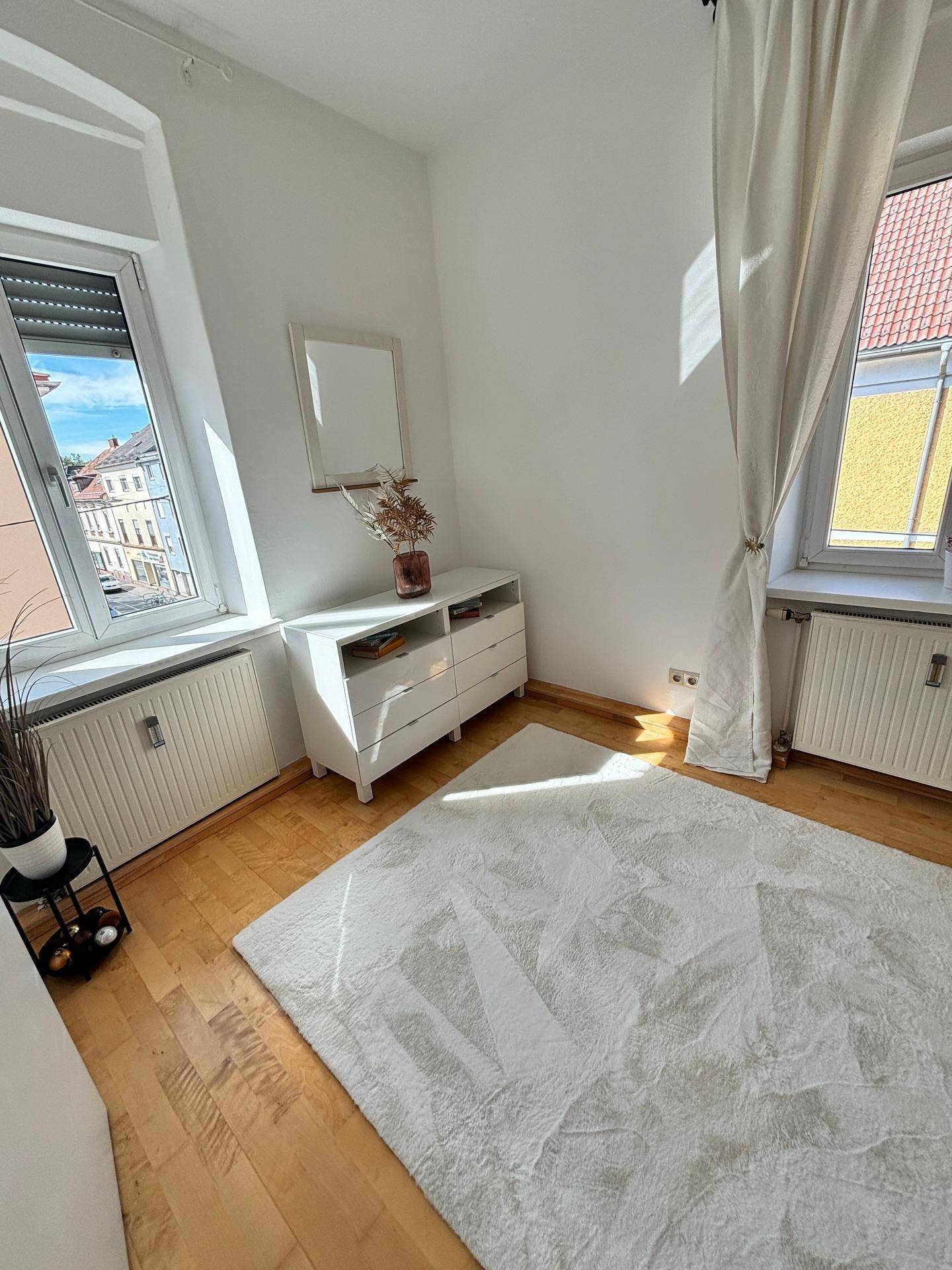 Helles Zimmer mit zwei Fenstern, Holzfußboden und weißer Kommode.
