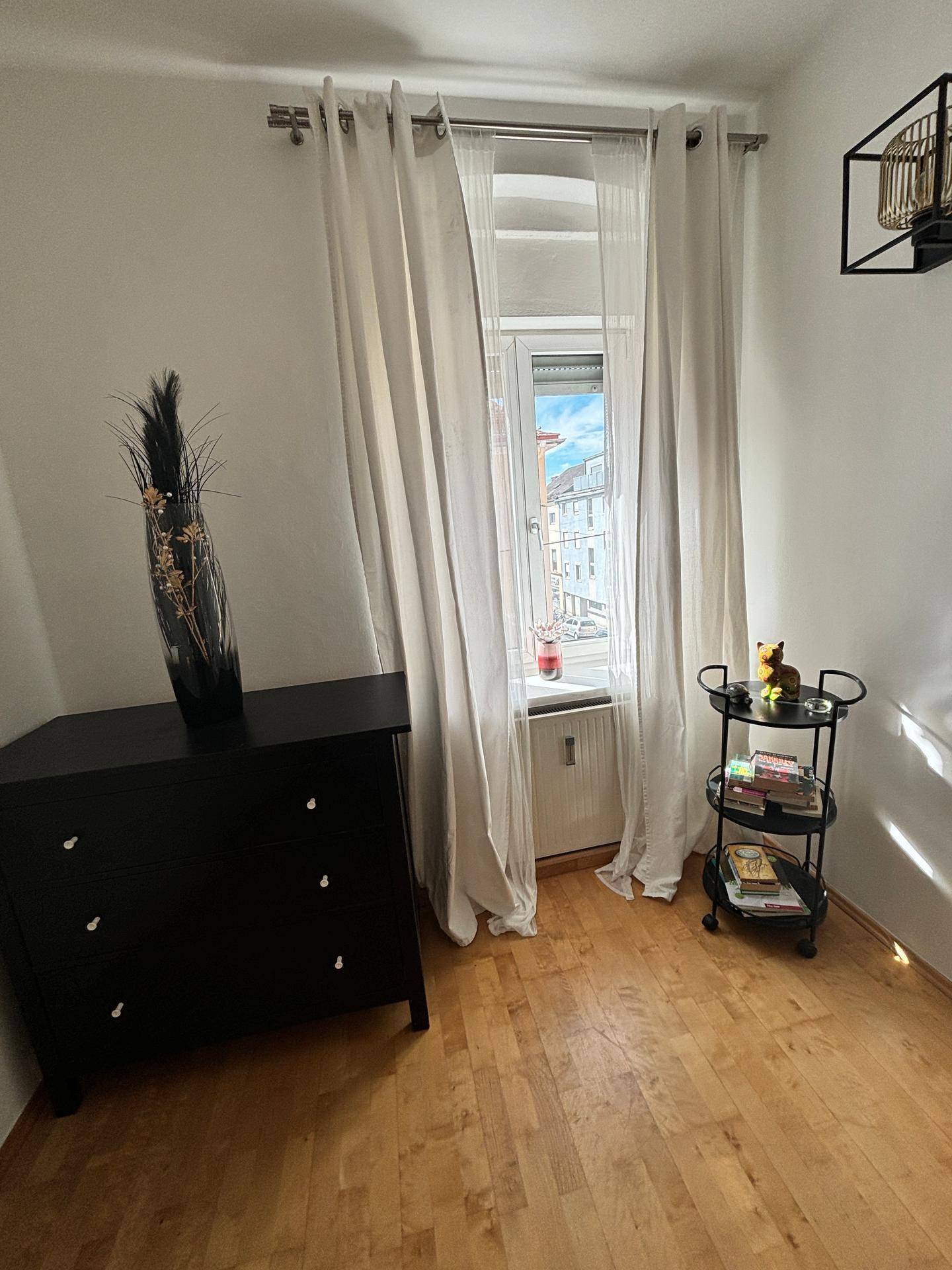 Helles Zimmer mit Holzfußboden, Fenster und einem Beistellwagen als Ablage.