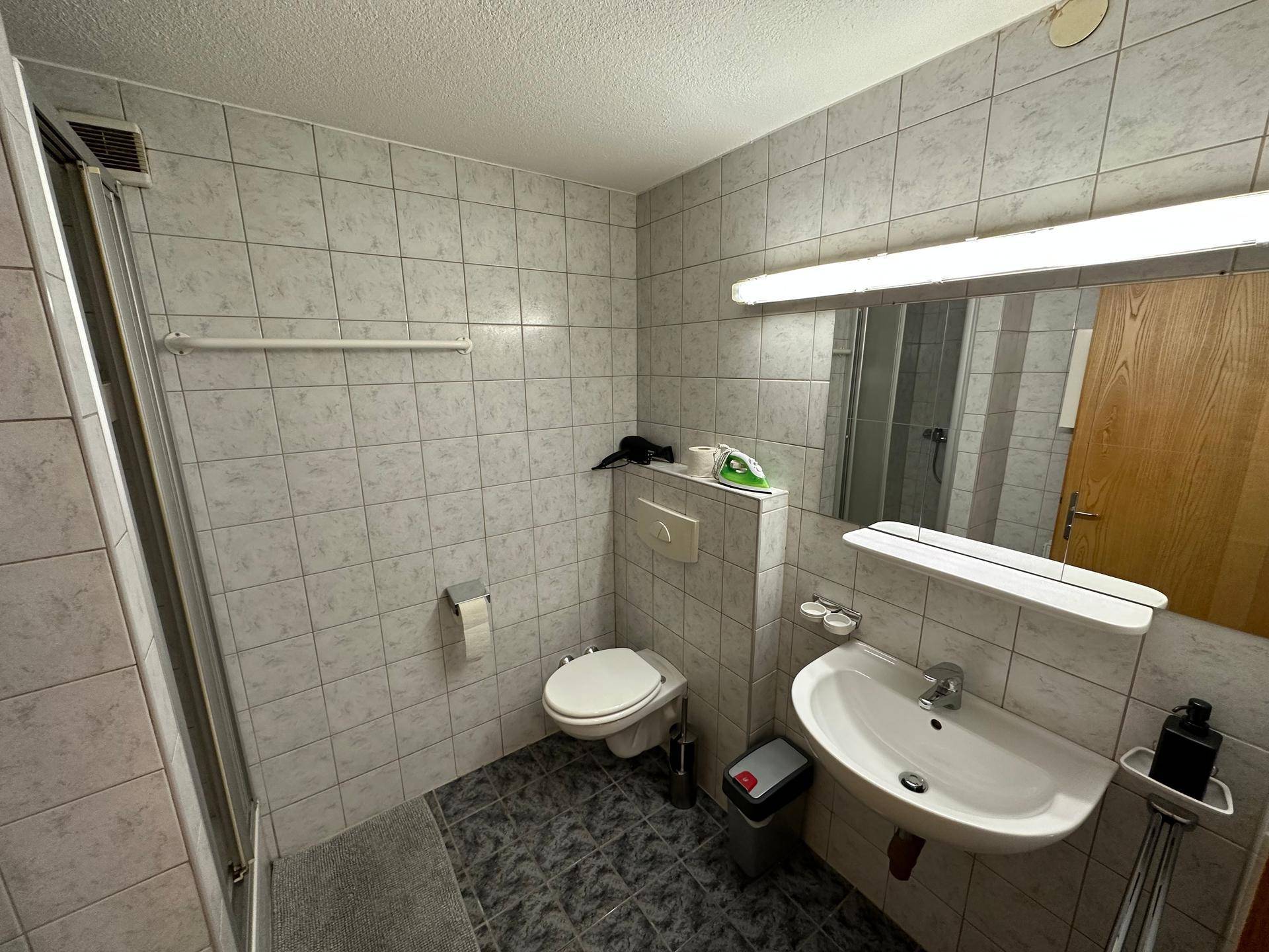 Badezimmer mit Waschbecken, Spiegel und Duschkabine, komplett gefliest.