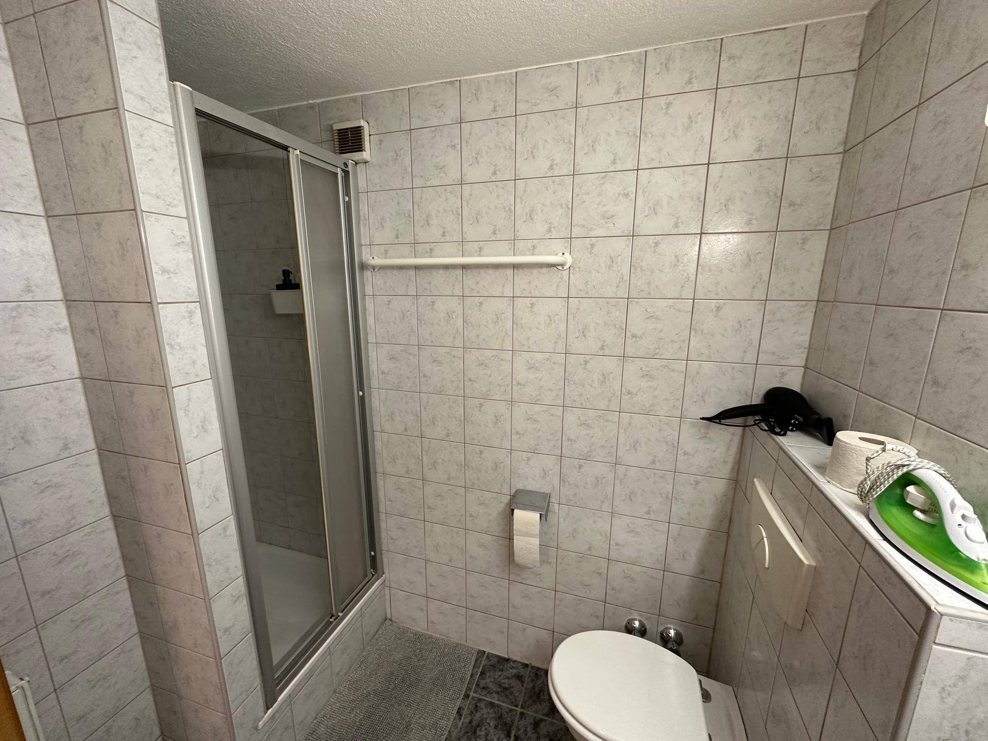 Gefliestes Badezimmer mit Duschkabine und wandhängendem WC in neutralen Farbtönen.