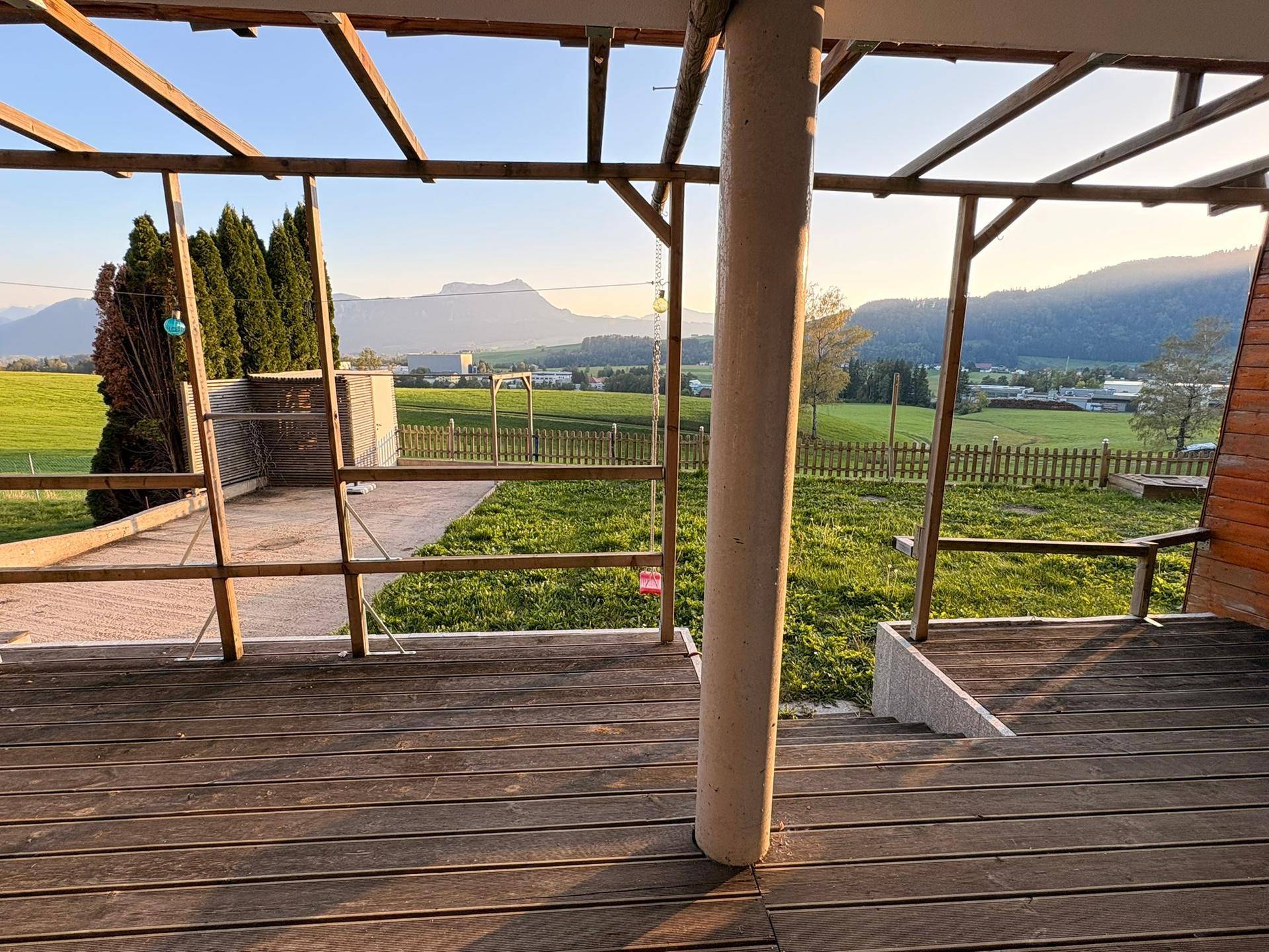 Holzterrasse mit Pergola-Struktur und weitem Blick auf grüne Felder und Berge.