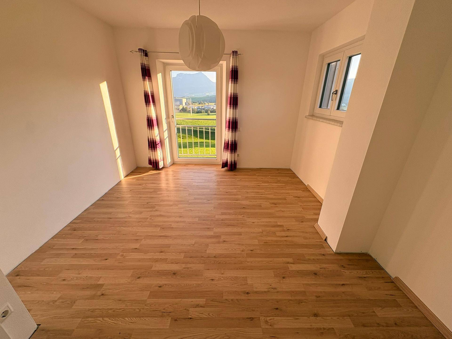 Lichtdurchflutetes Zimmer mit Laminatboden, Vorhängen und Zugang zu einem französischen Balkon.