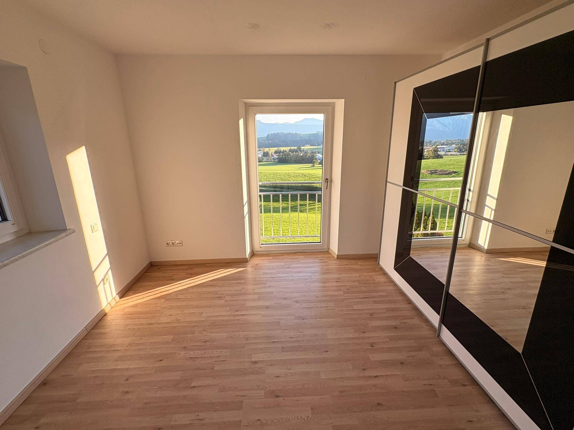 Helles Zimmer mit Laminatboden, großem Fenster und Zugang zu einem französischen Balkon.