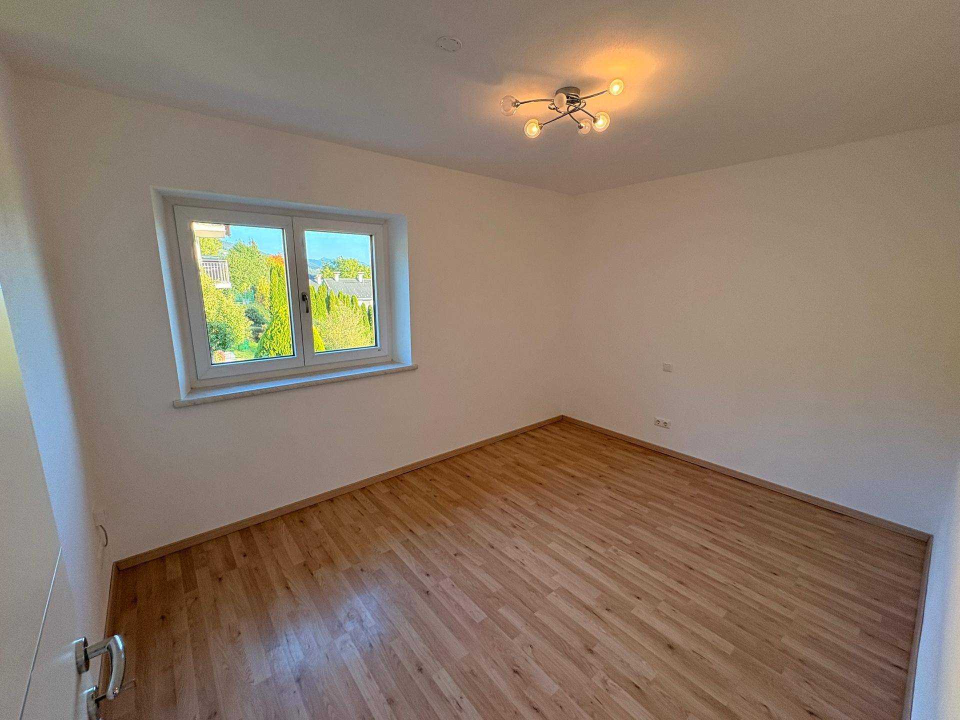 Helles, leeres Zimmer mit Laminatboden und einem Fenster, das viel Tageslicht hereinlässt.